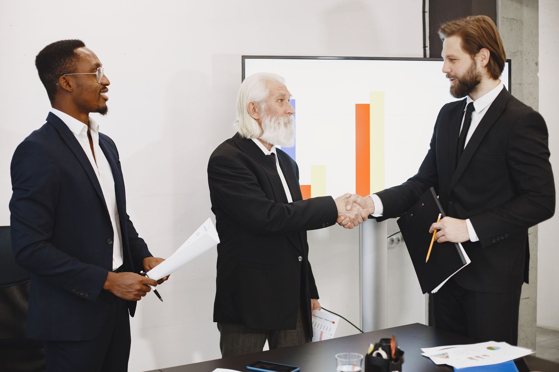 Three professionals in suits shake hands in a bright office meeting room.