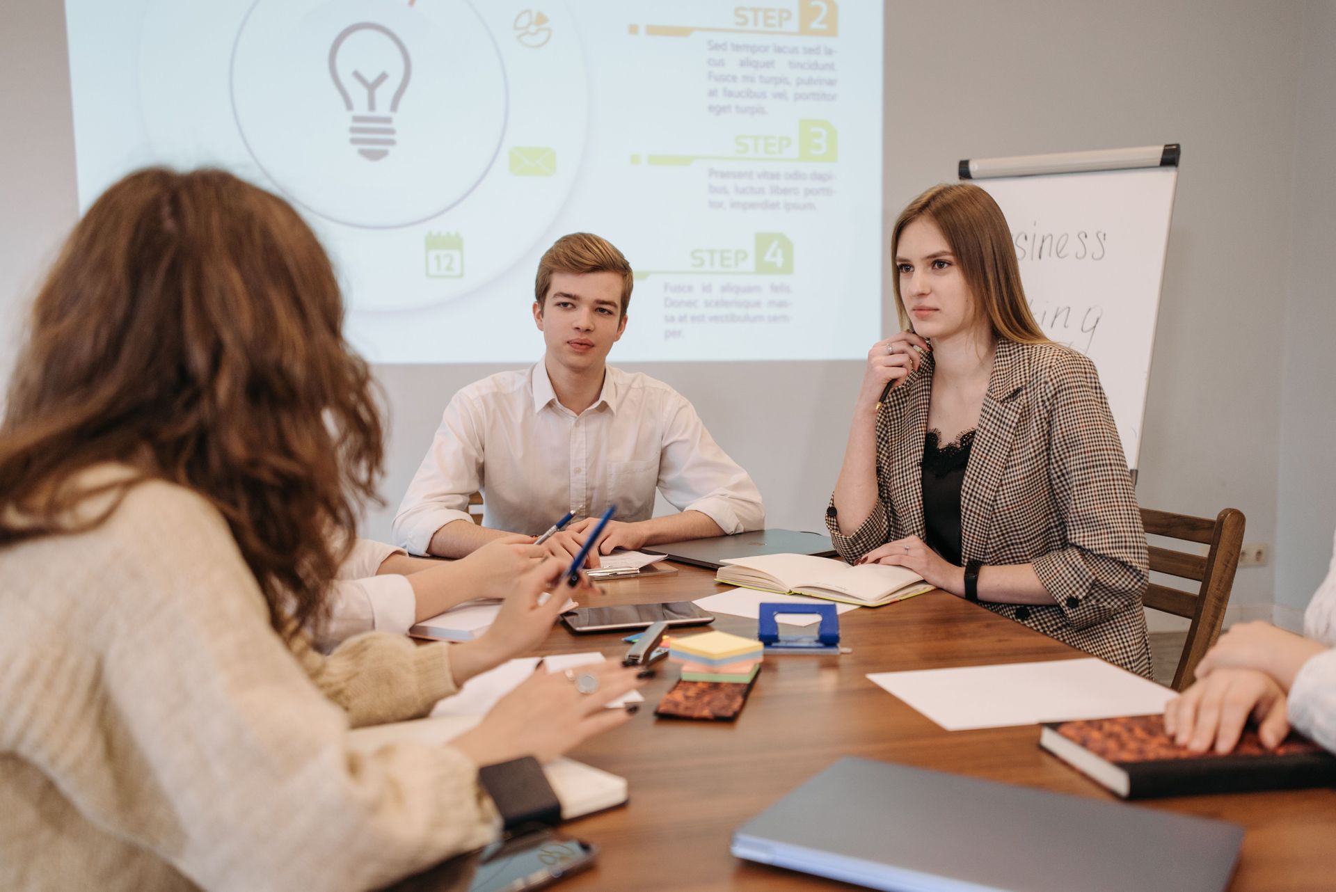 Business meeting around a conference table with documents, laptop, and presentation screen in the background