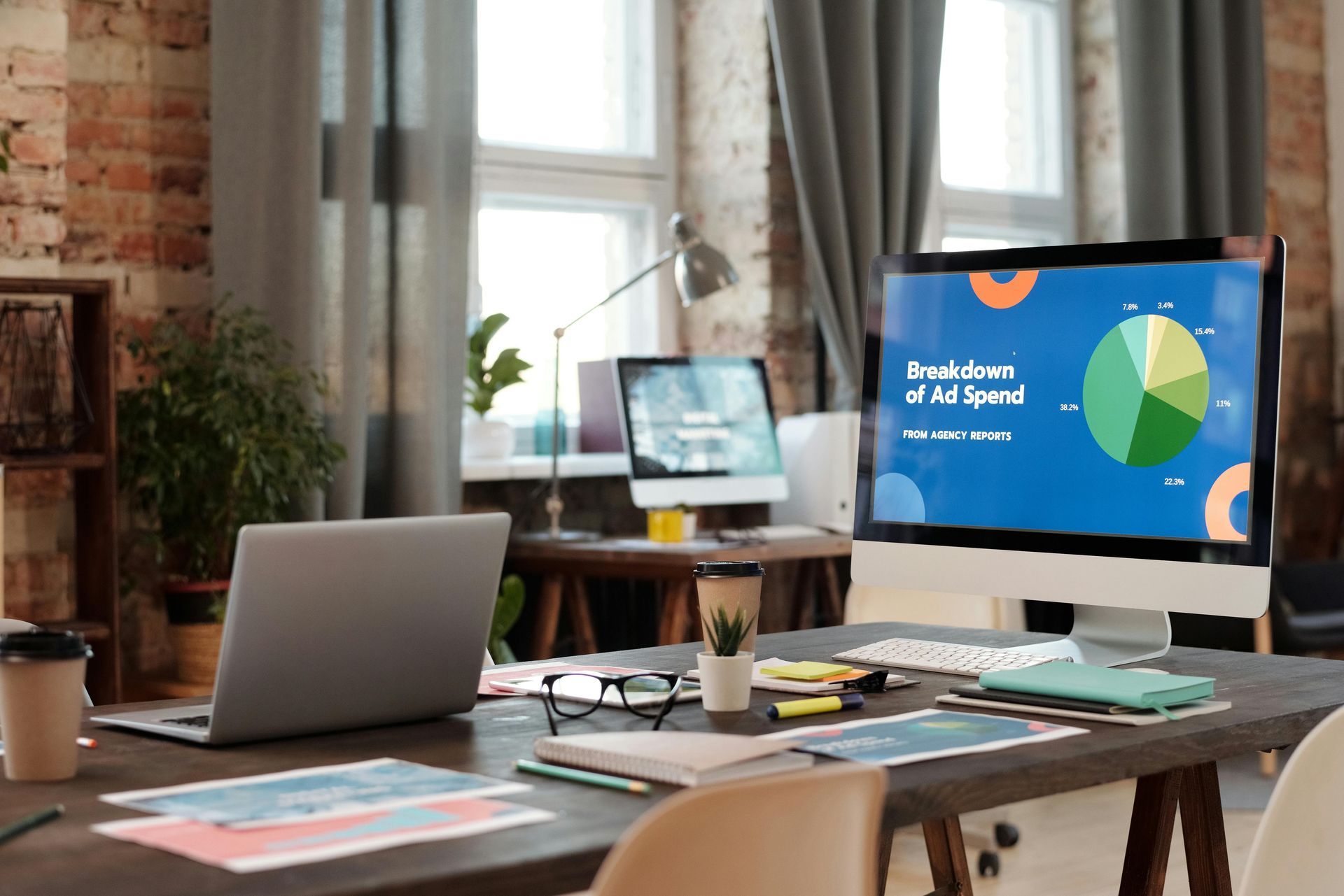 A desk with a laptop and computer monitor displaying an ad spend pie chart in a sunlit home office.