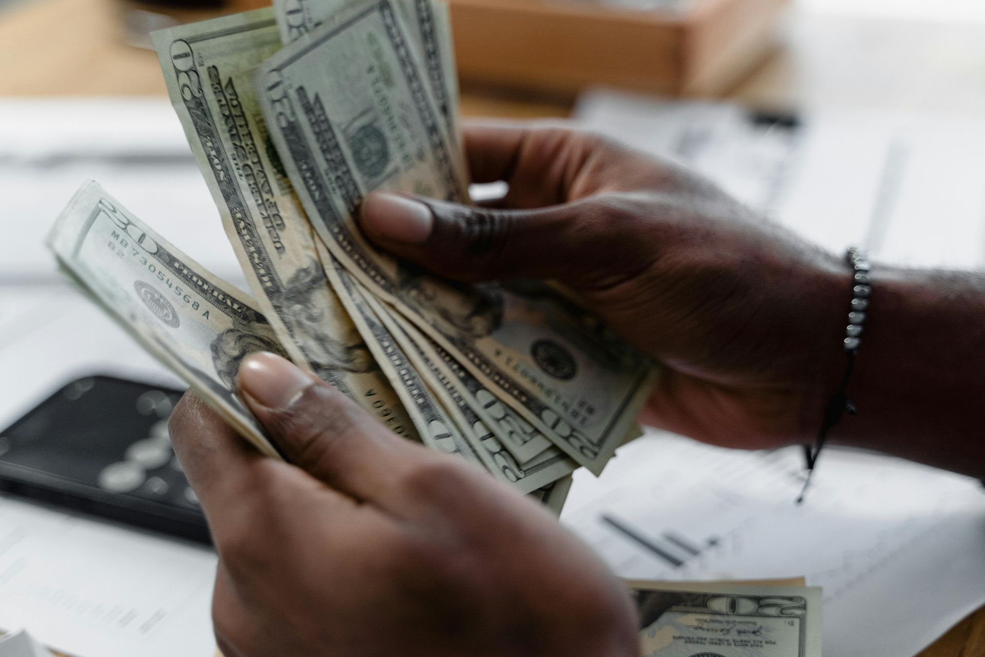 Hands holding a fan of U.S. twenty-dollar bills above a desk with documents and a smartphone.