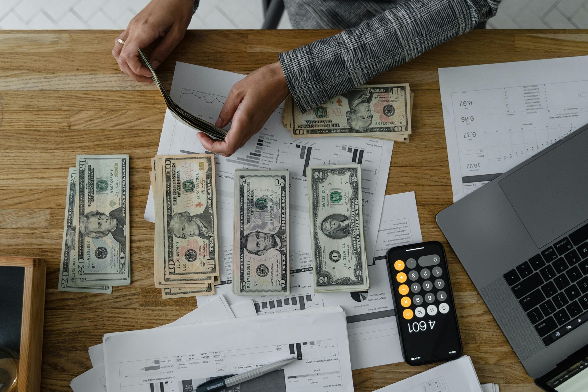 A person works at a desk, counting and organizing several stacks of US dollar bills near a calculator and laptop.