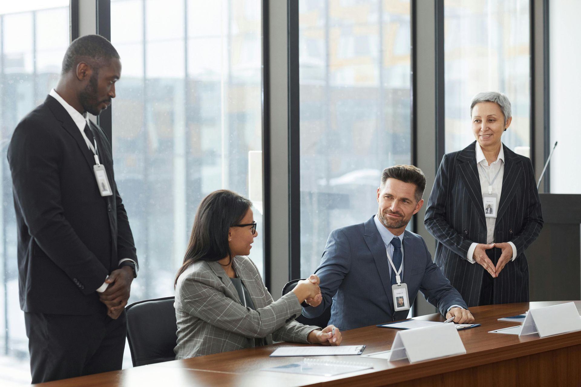 A woman in a blazer shakes hands with a man in a suit across a conference table; two colleagues stand nearby in an office.