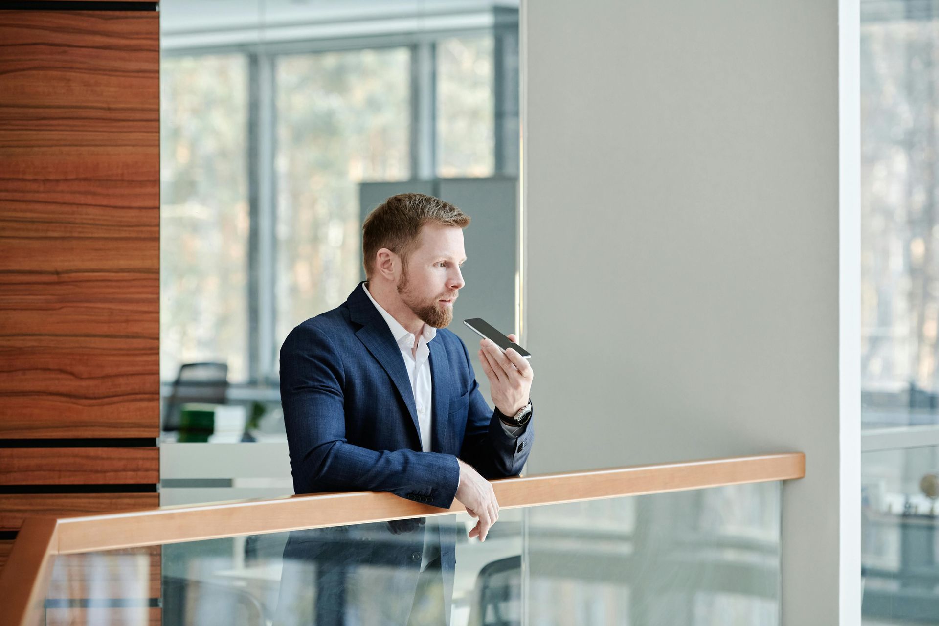 Man in a navy suit speaking into a microphone on a balcony in a bright office lobby