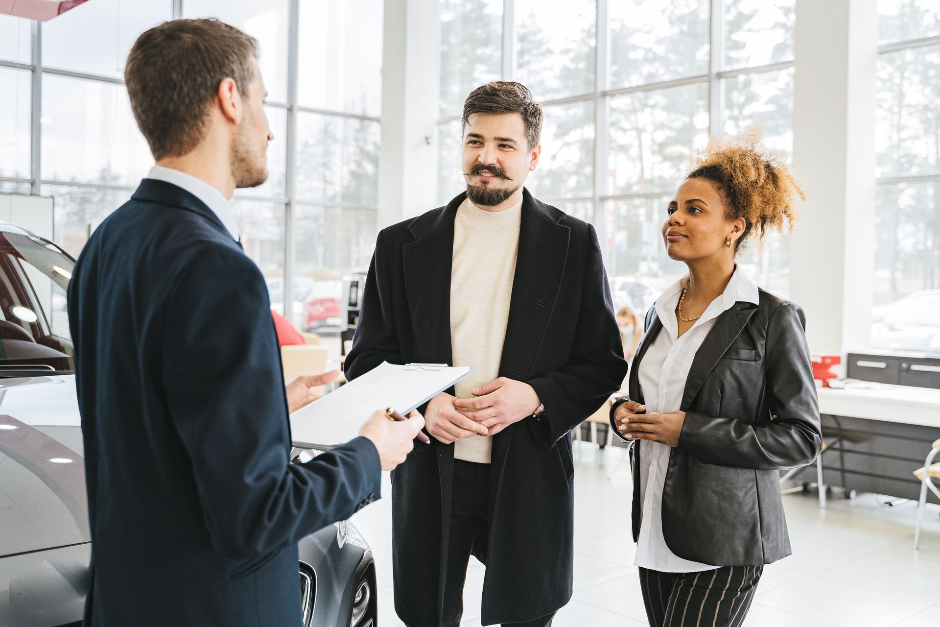 Three people talking in a bright car showroom, with a salesperson holding paperwork beside a car.