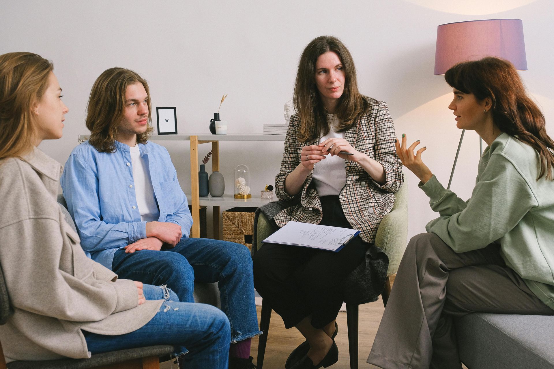 Four people in a circle, one speaking while others listen. A therapist gestures with a pen.
