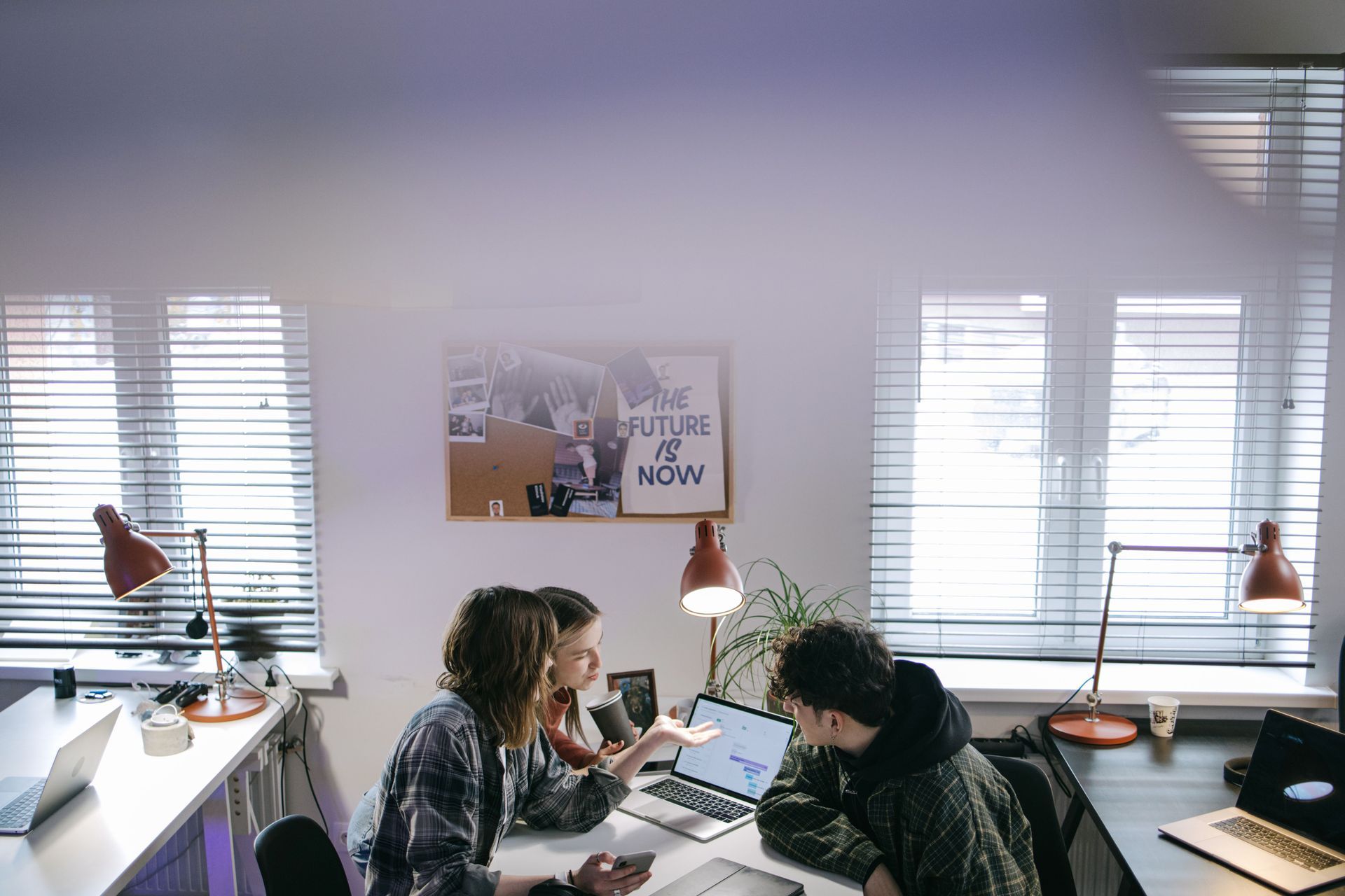 Two people working at a desk in a bright office with laptops and desk lamps