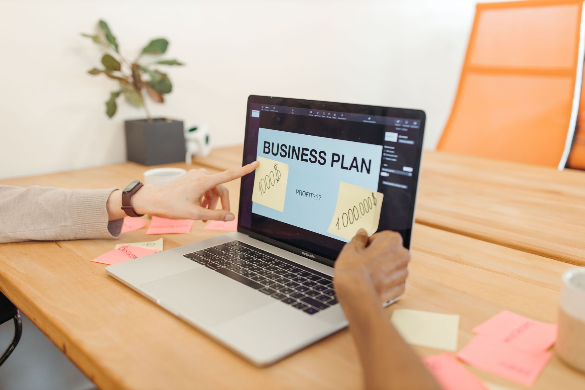 Hands reviewing a laptop displaying a business plan on a wooden desk with sticky notes