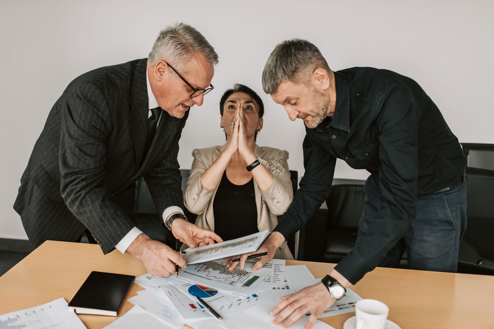 Three people review documents at a table, one appearing stressed while two lean in to discuss.
