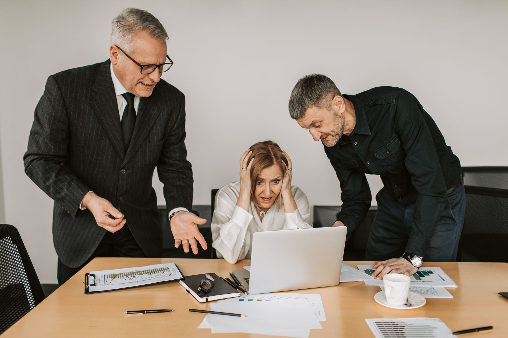 Three coworkers gathered around a laptop in an office, looking concerned and discussing documents.