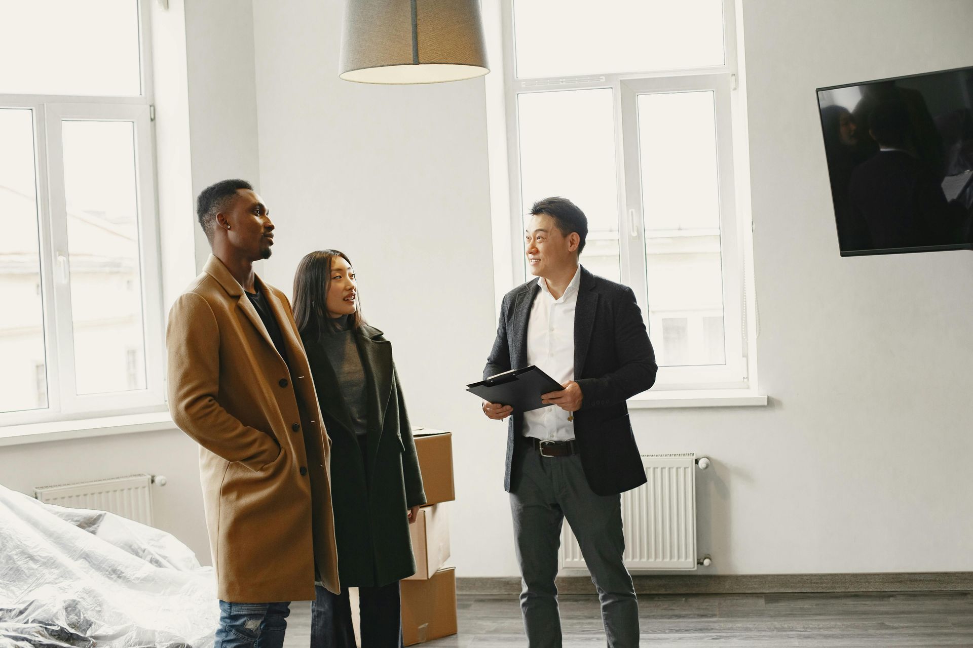 A professional in a suit holds a clipboard, talking to two people standing in a bright, sparsely furnished room.
