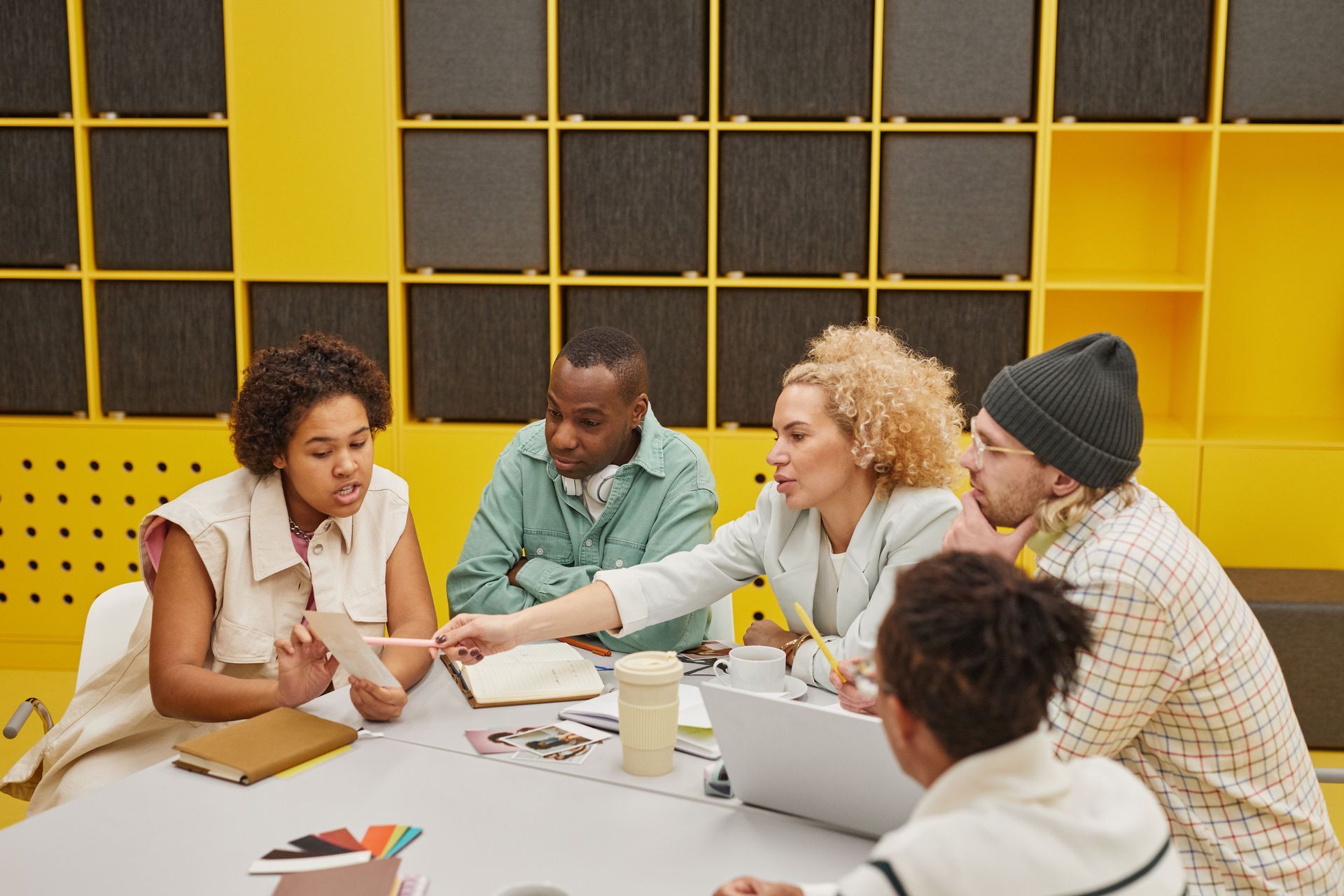 Five people collaborate around a table in a yellow meeting room with laptops, notebooks, and coffee.