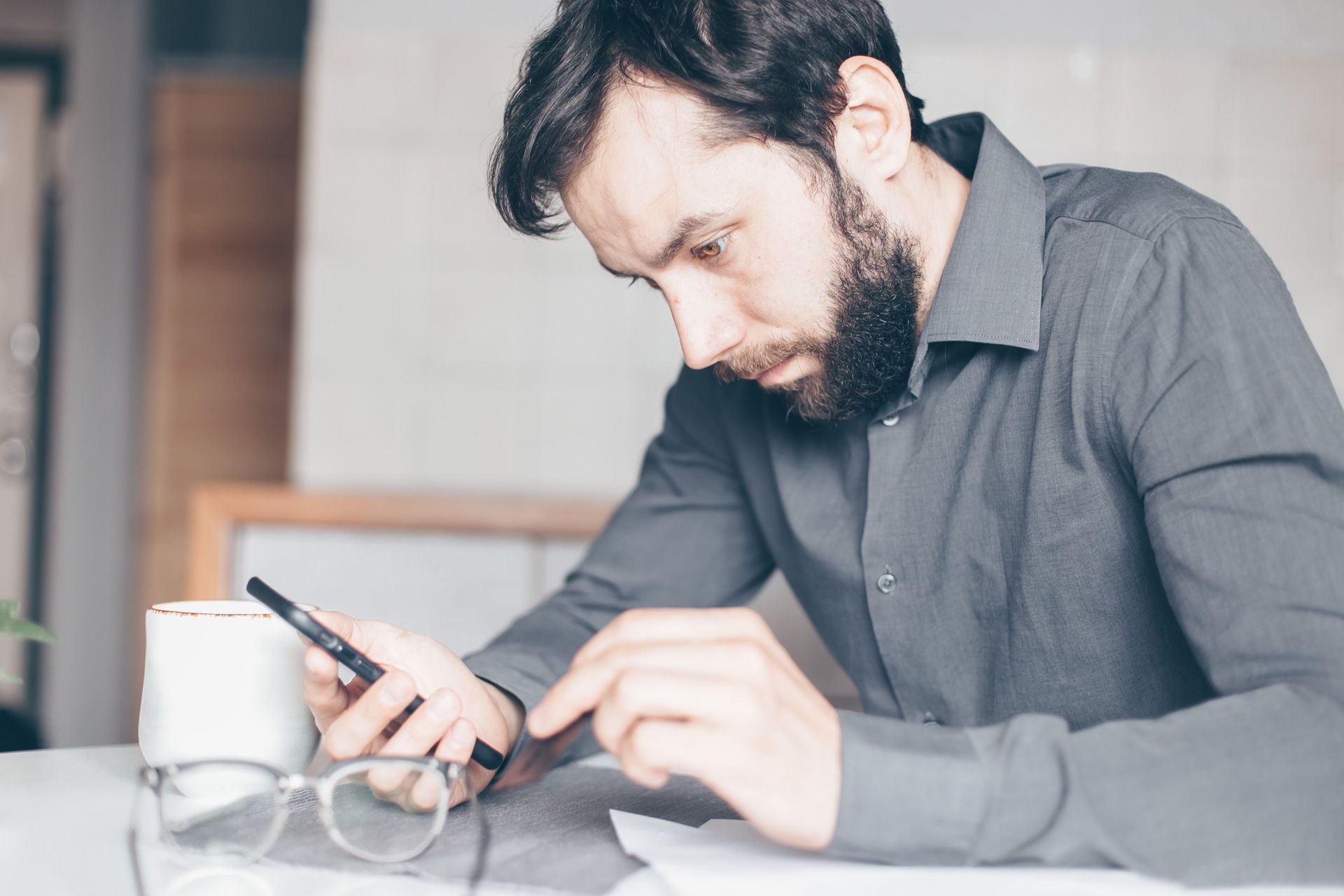 Man in a gray shirt using a smartphone at a desk with a mug and eyeglasses