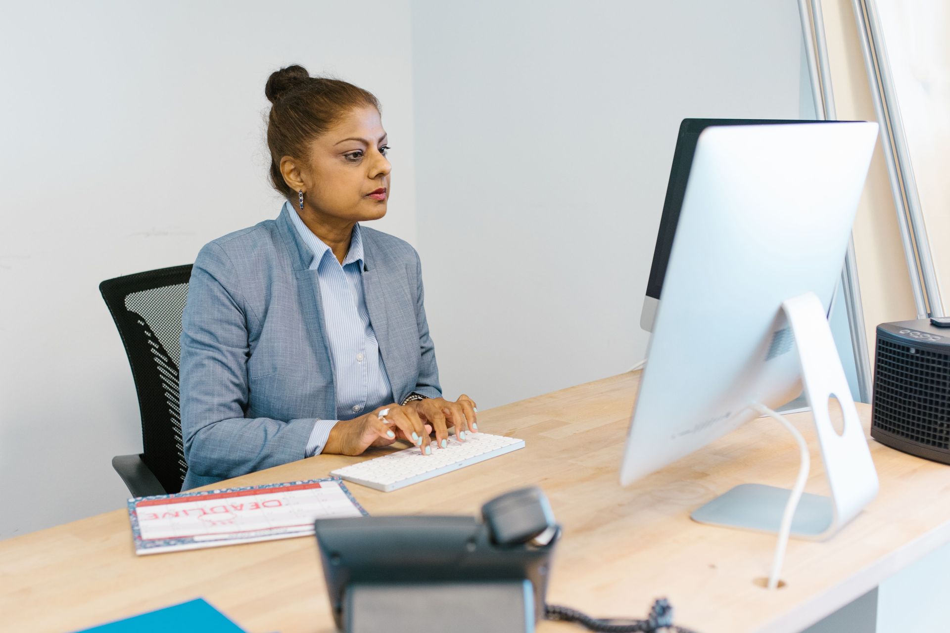Woman seated at a desk, looking at a desktop monitor in a bright office