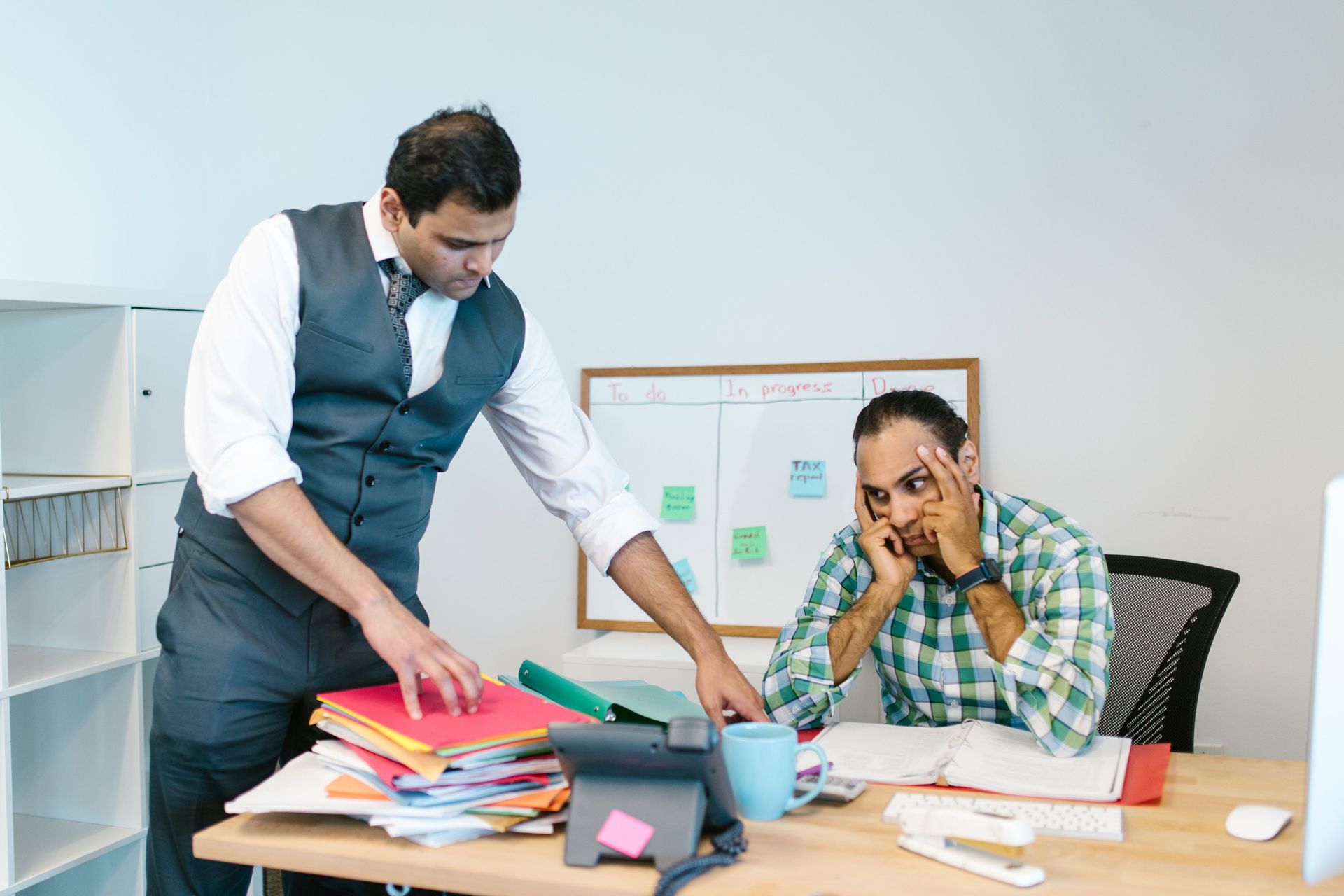Two coworkers in an office, one standing over a cluttered desk while the other looks stressed on the phone.