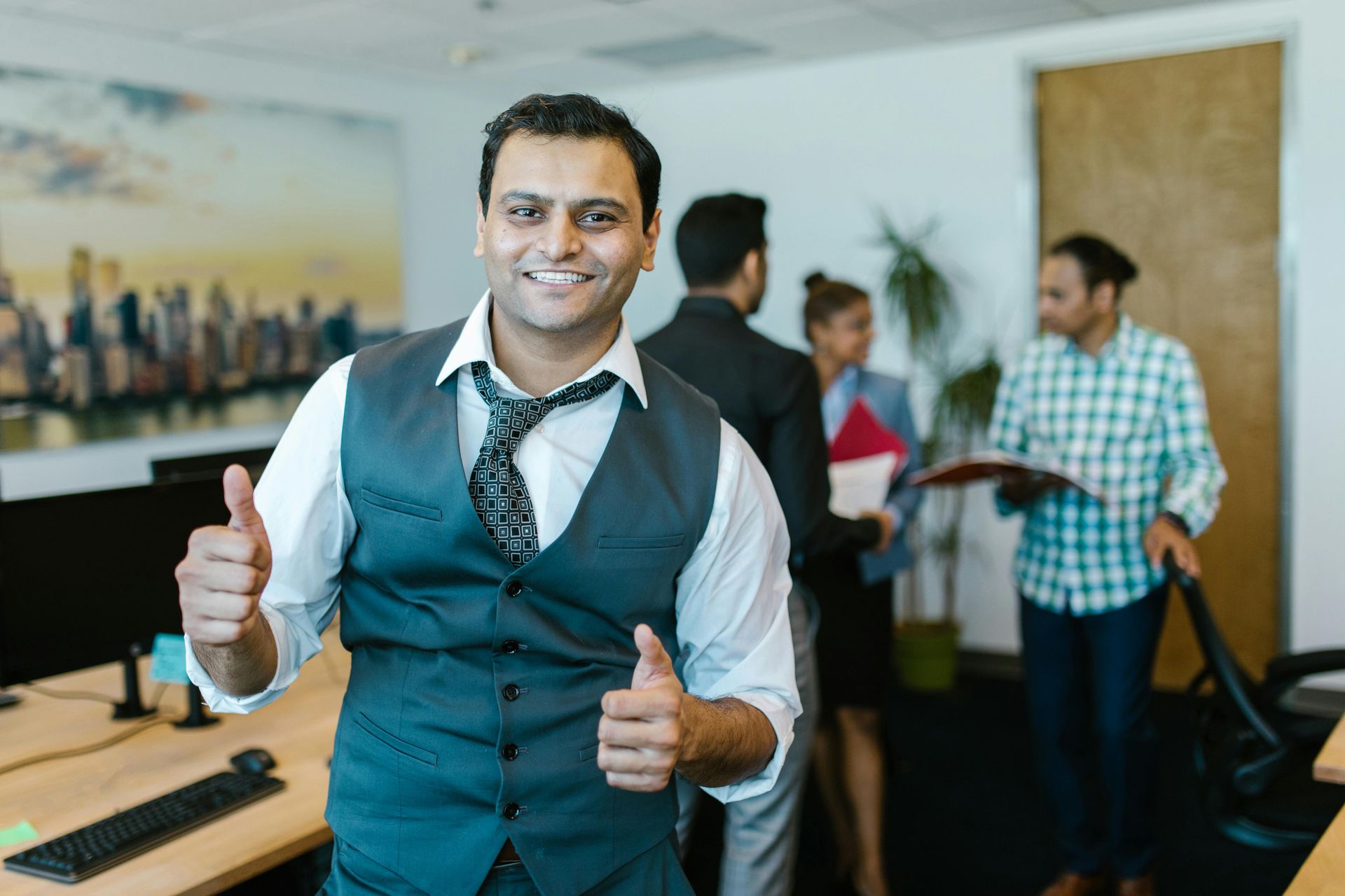 A smiling professional in a vest and tie gives a thumbs-up in an office with coworkers standing in the background.