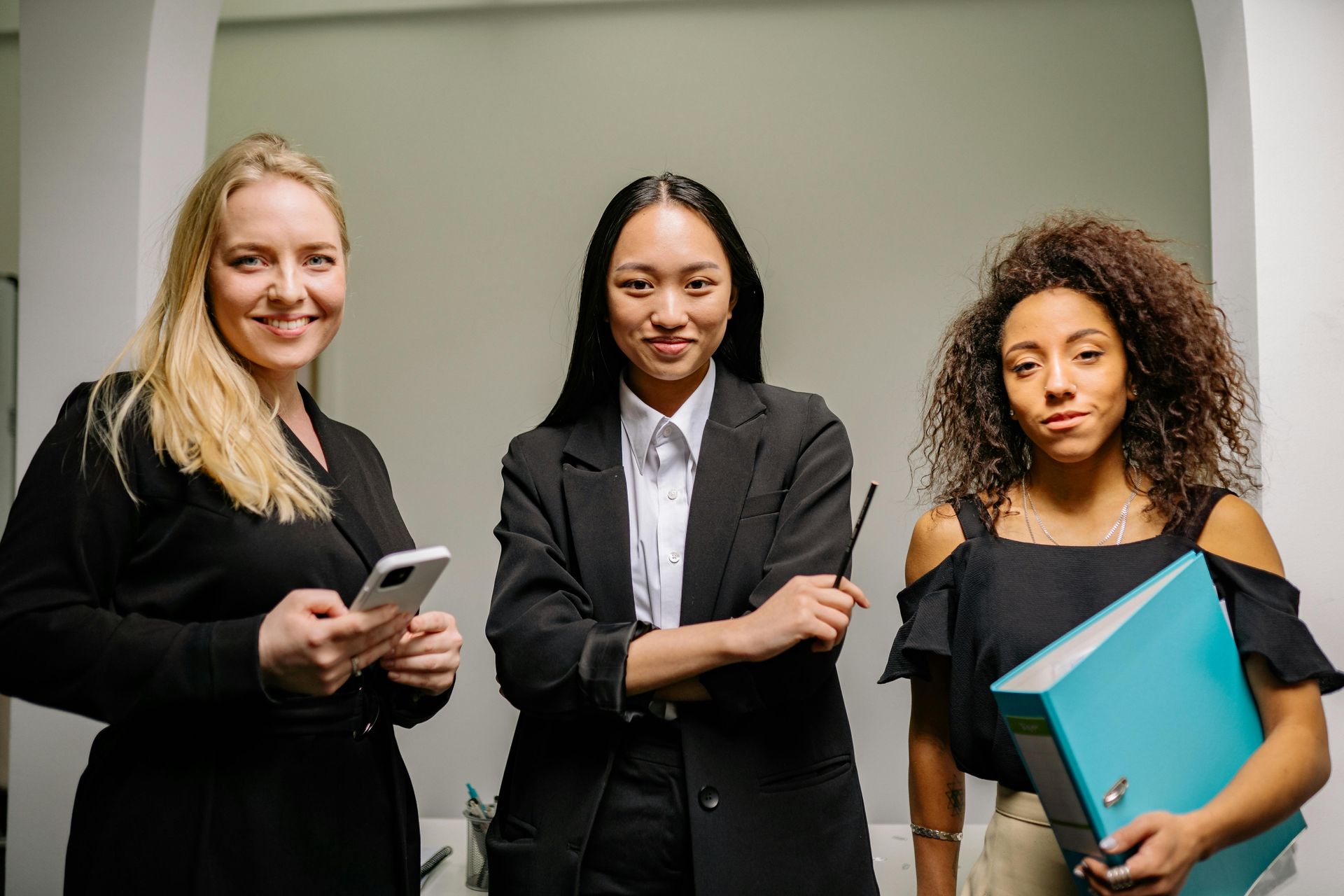 Three professionals stand together in an office; one holds a smartphone, one a pen, and one a teal binder.
