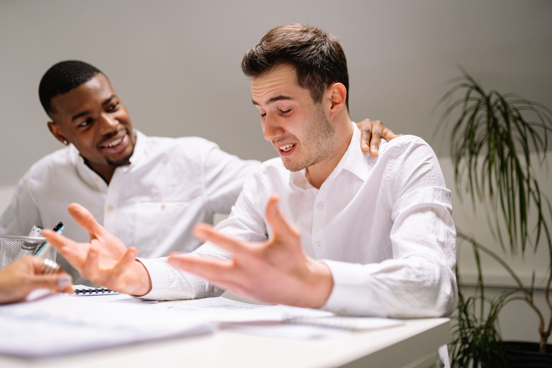 Two coworkers in white shirts discussing something at a desk, one gesturing with open hands.