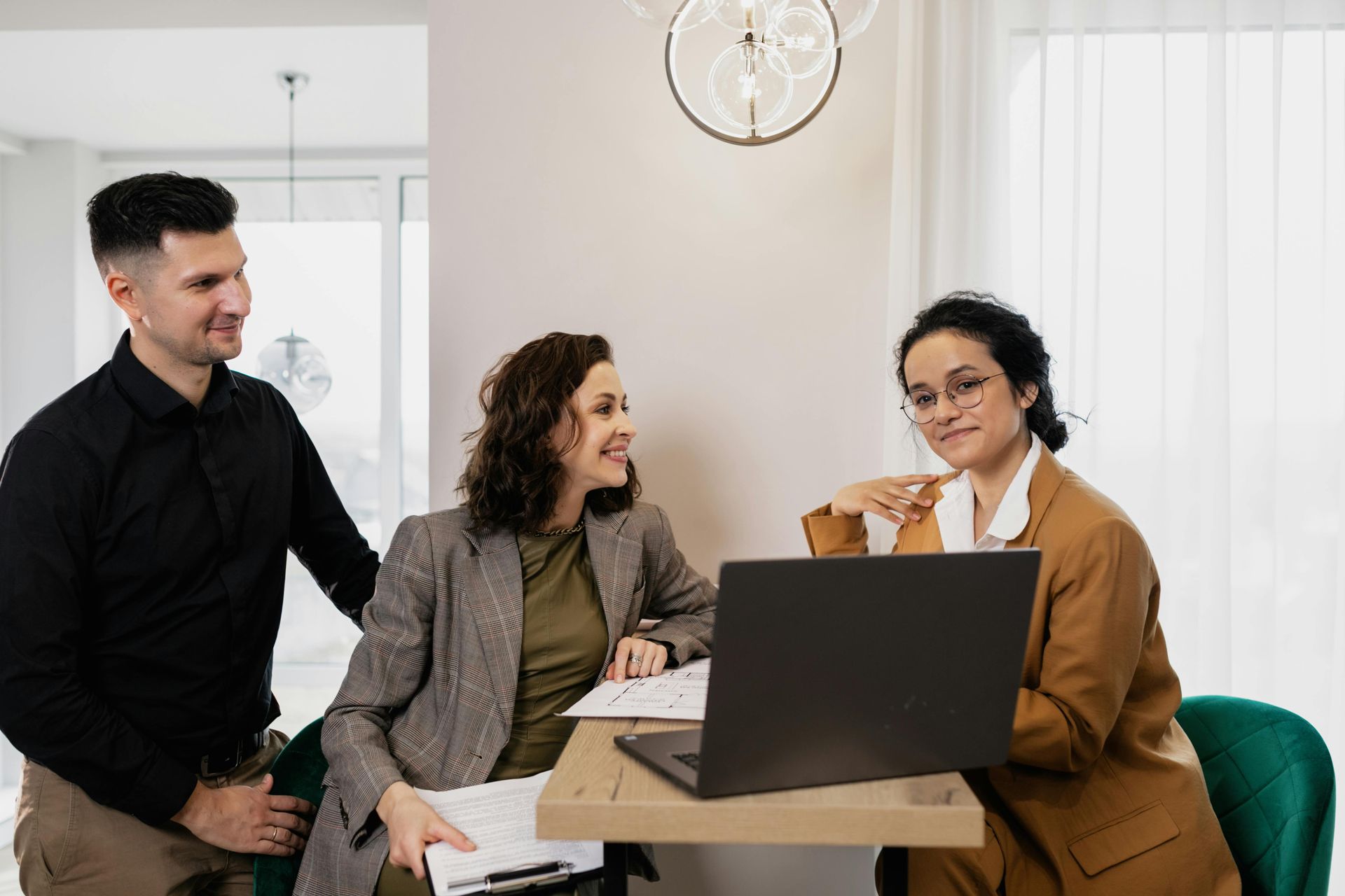 Three professionals in business attire collaborate around a laptop on a small table in a bright, modern office space.