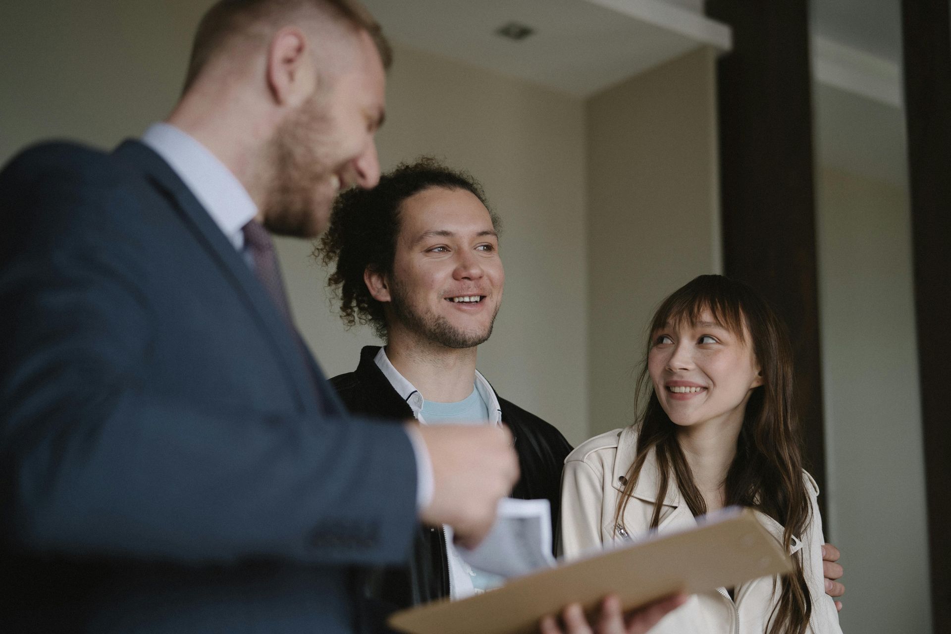 A professional in a suit presents a folder to a couple who are looking at him with smiles in a well-lit indoor space.