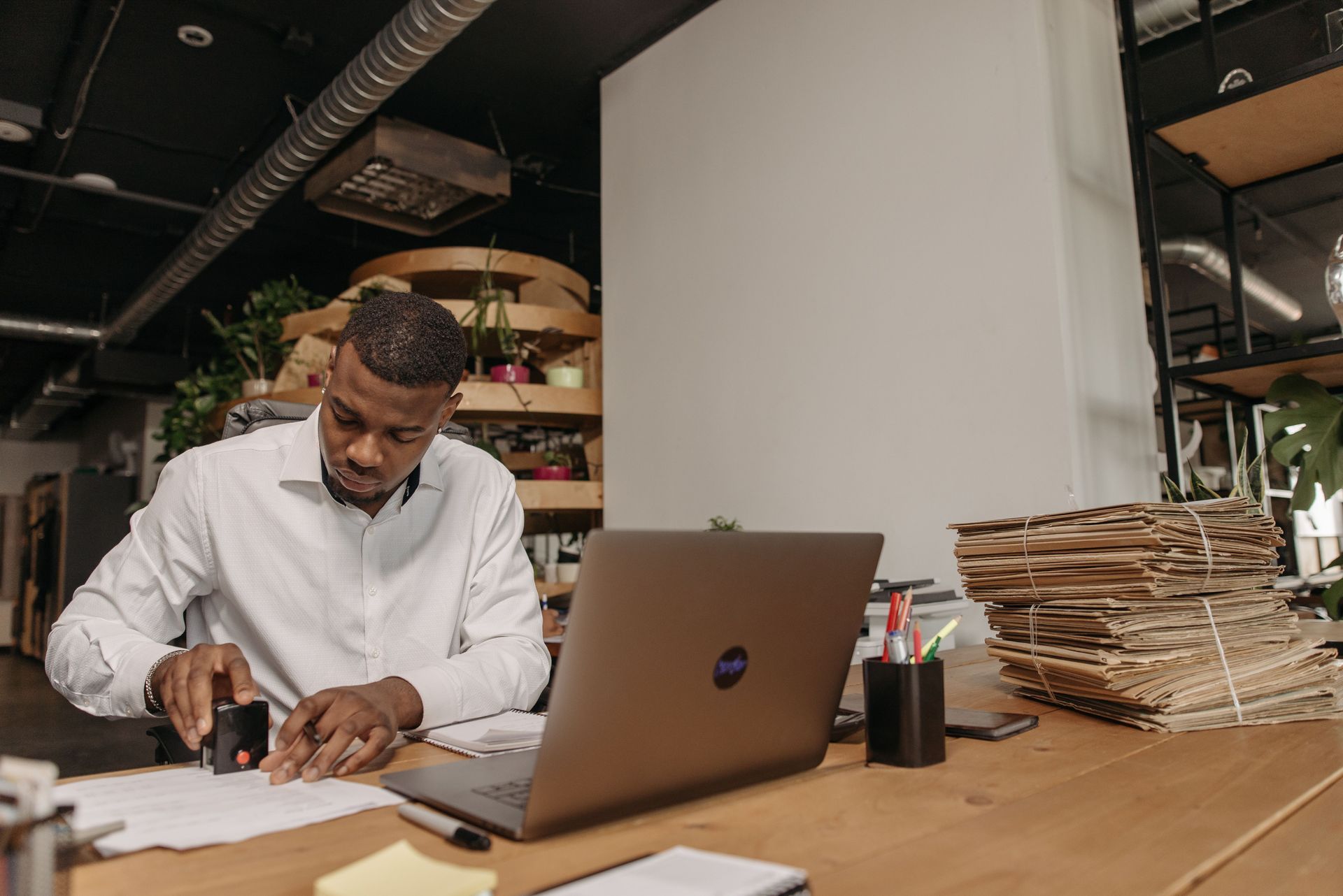 Man in white shirt working at a desk in a modern office, writing beside a laptop and stacked papers