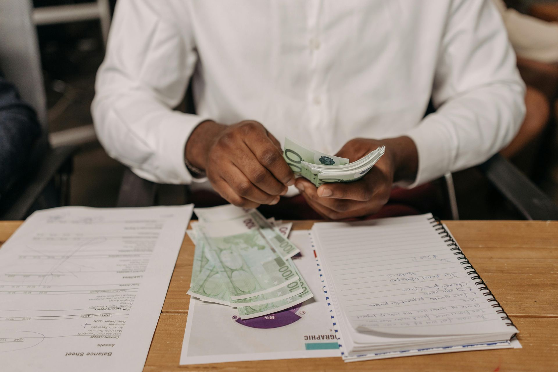 A person in a white shirt counting a stack of green banknotes at a wooden desk with papers and a notebook.