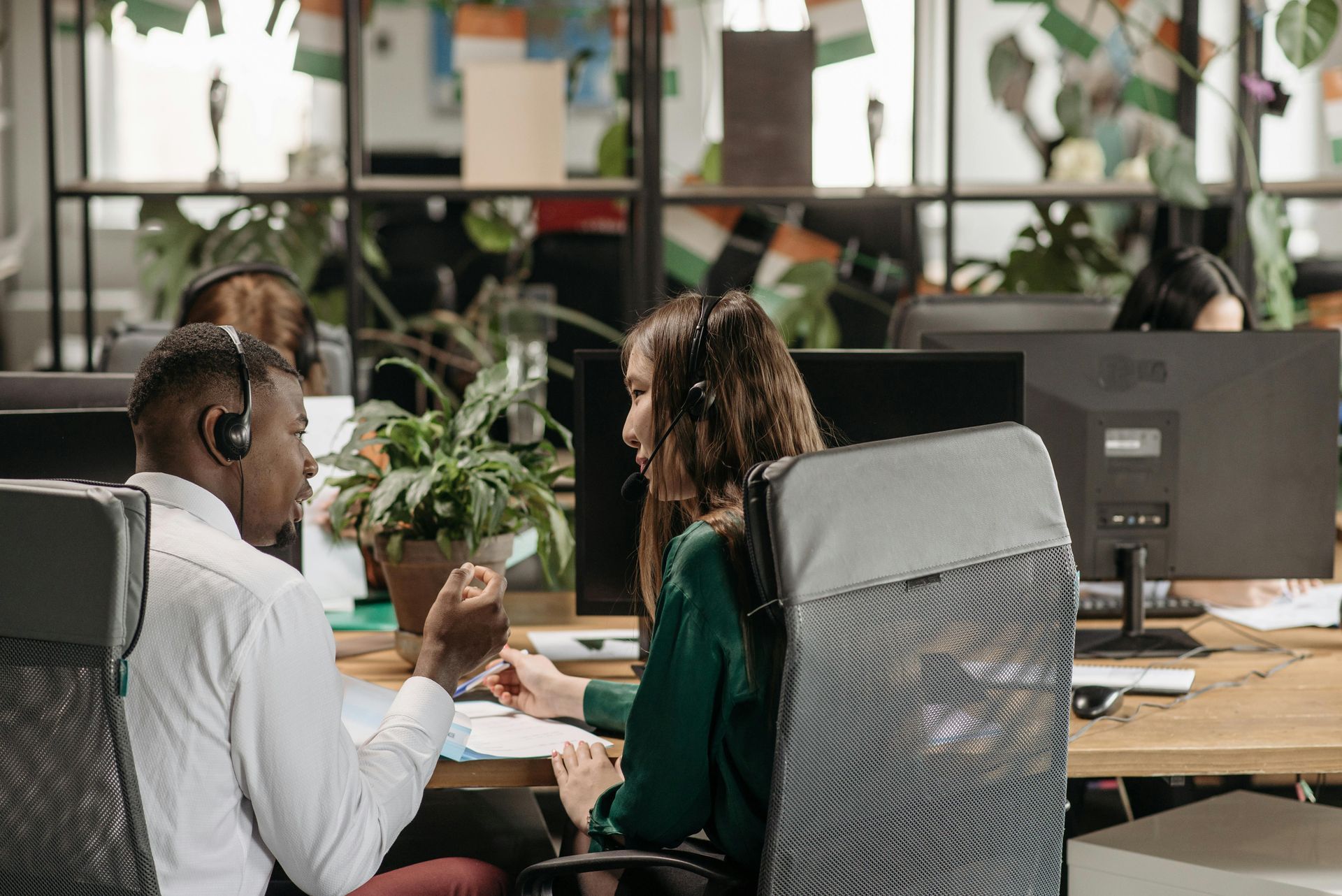 Two people with headsets at desks in an office; they're looking at papers and conversing, surrounded by computers and plants.