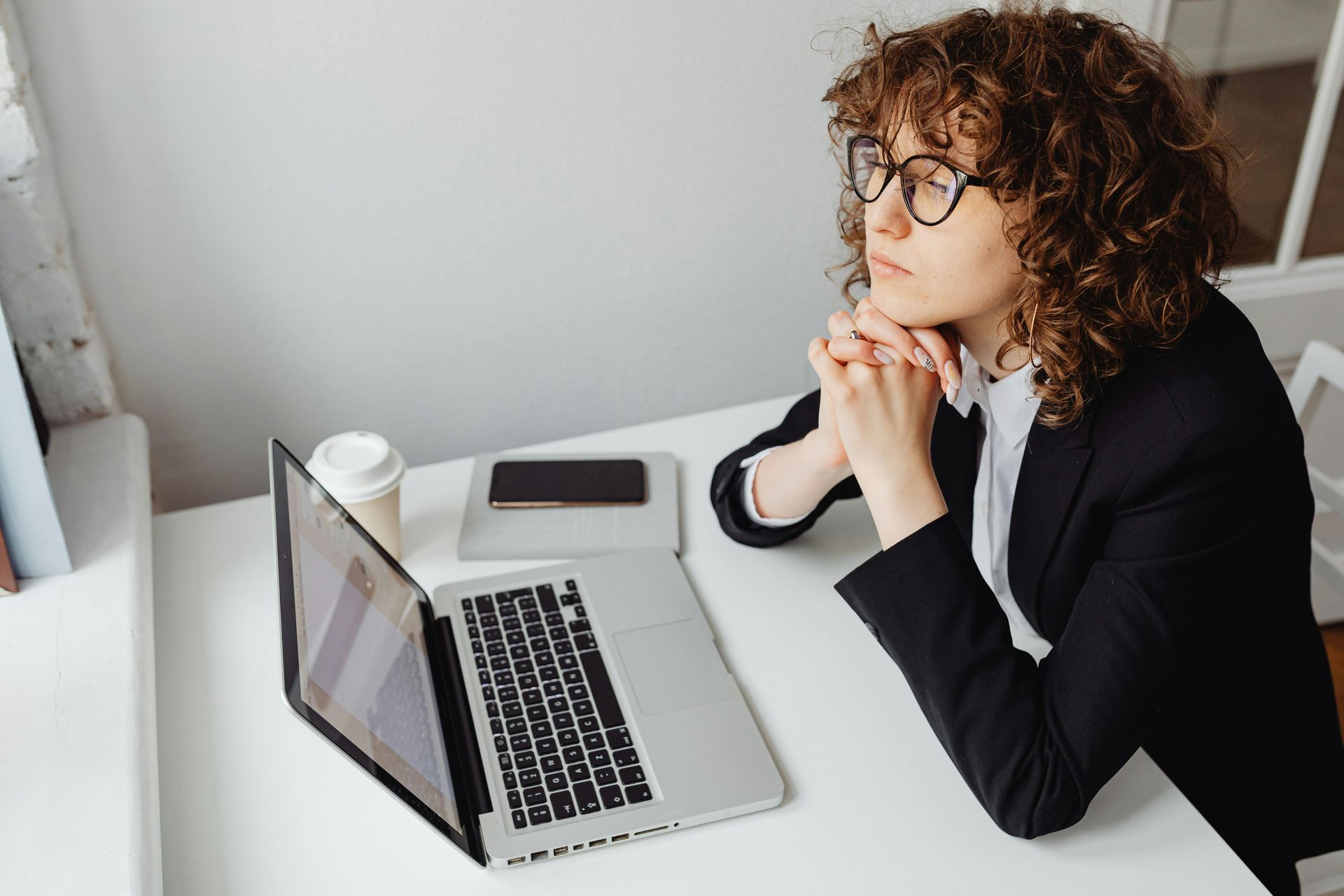 A person with curly hair and glasses sits at a white desk with a laptop and coffee, hands clasped, looking thoughtful.