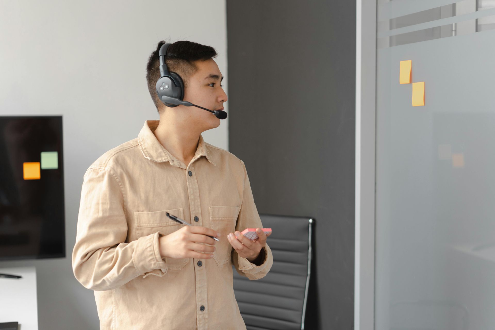 Man with headset gestures near a whiteboard with sticky notes in a modern office setting.
