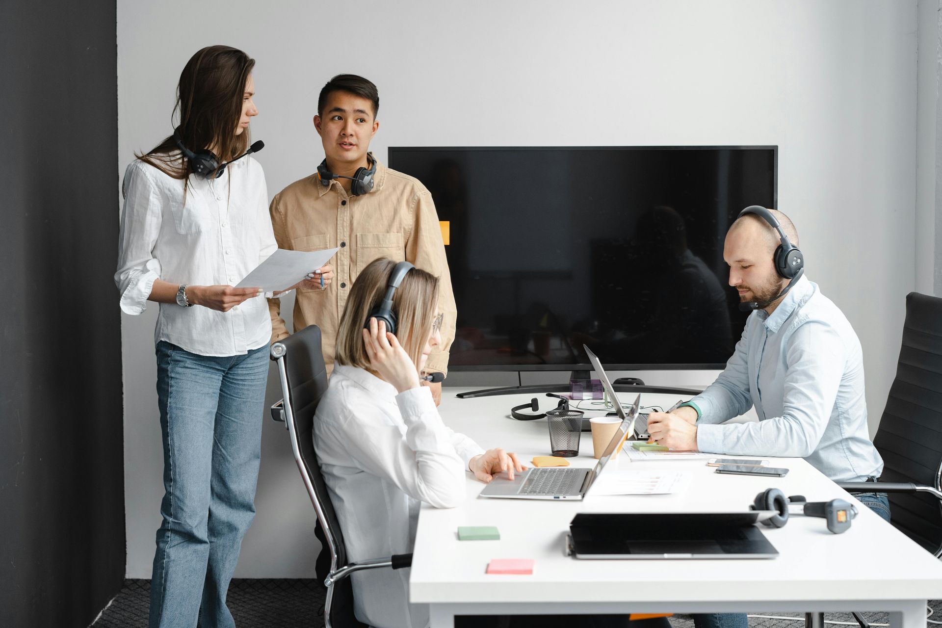 Four people wearing headsets in an office; two standing and two seated at a desk with laptops.