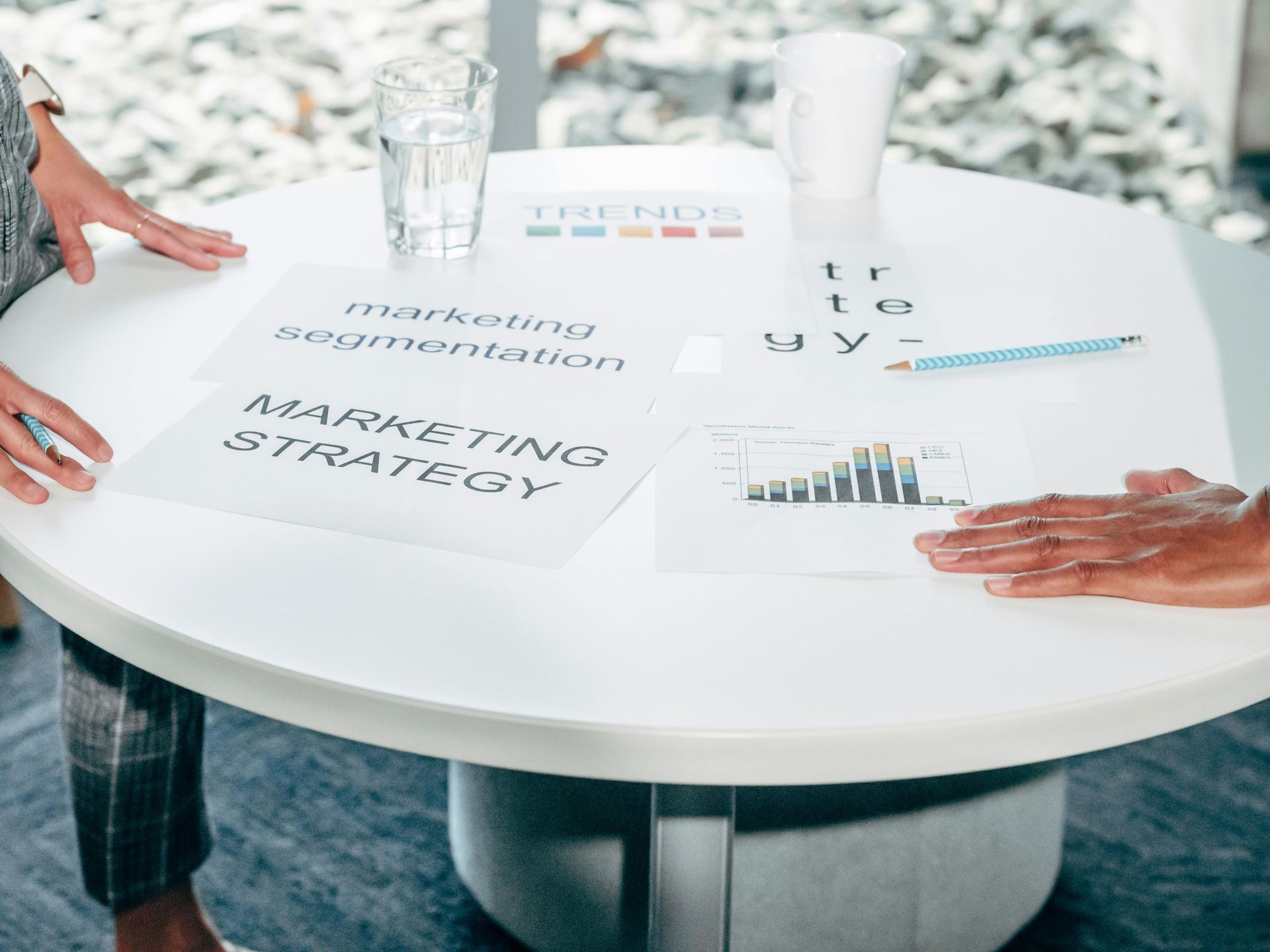 Hands around a white table with marketing strategy notes, charts, and glasses in a bright office setting
