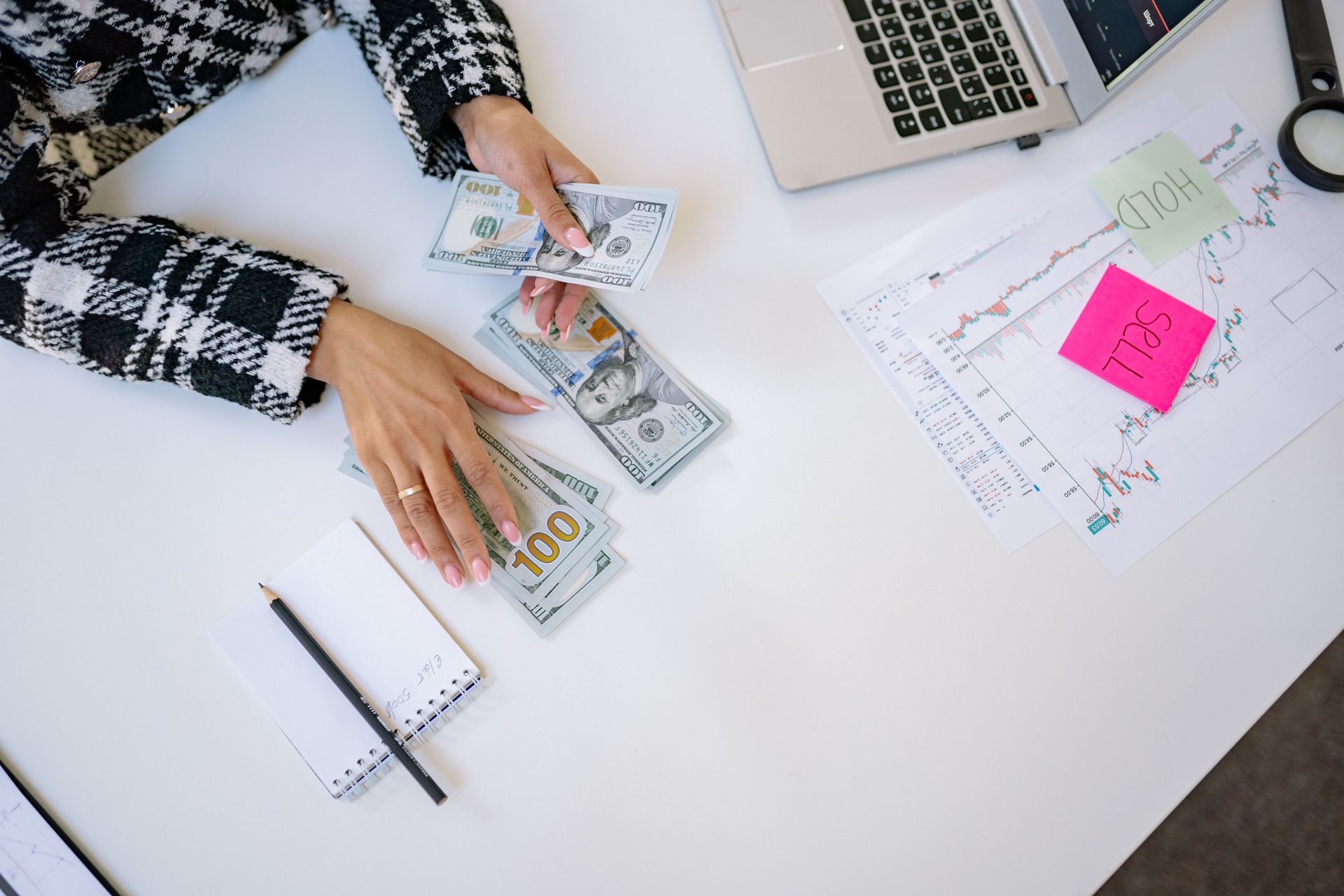 Hands counting cash at a desk with a laptop, notebook, and financial charts with sticky notes.