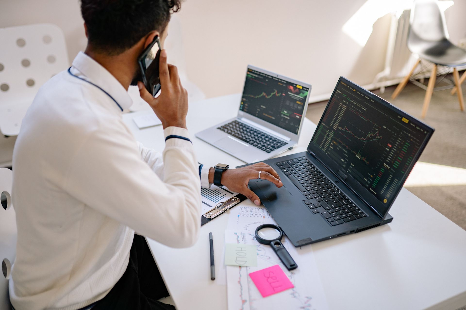 Person on a phone at a desk with dual laptops displaying code in a bright office