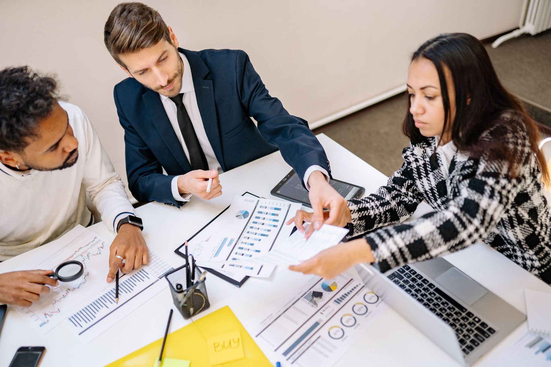 Three coworkers review charts around a table with laptop, documents, and magnifying glass in a meeting room