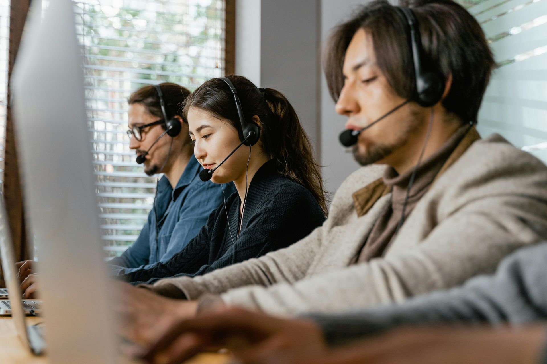 Three people wearing headsets sit at computers in an office, working.