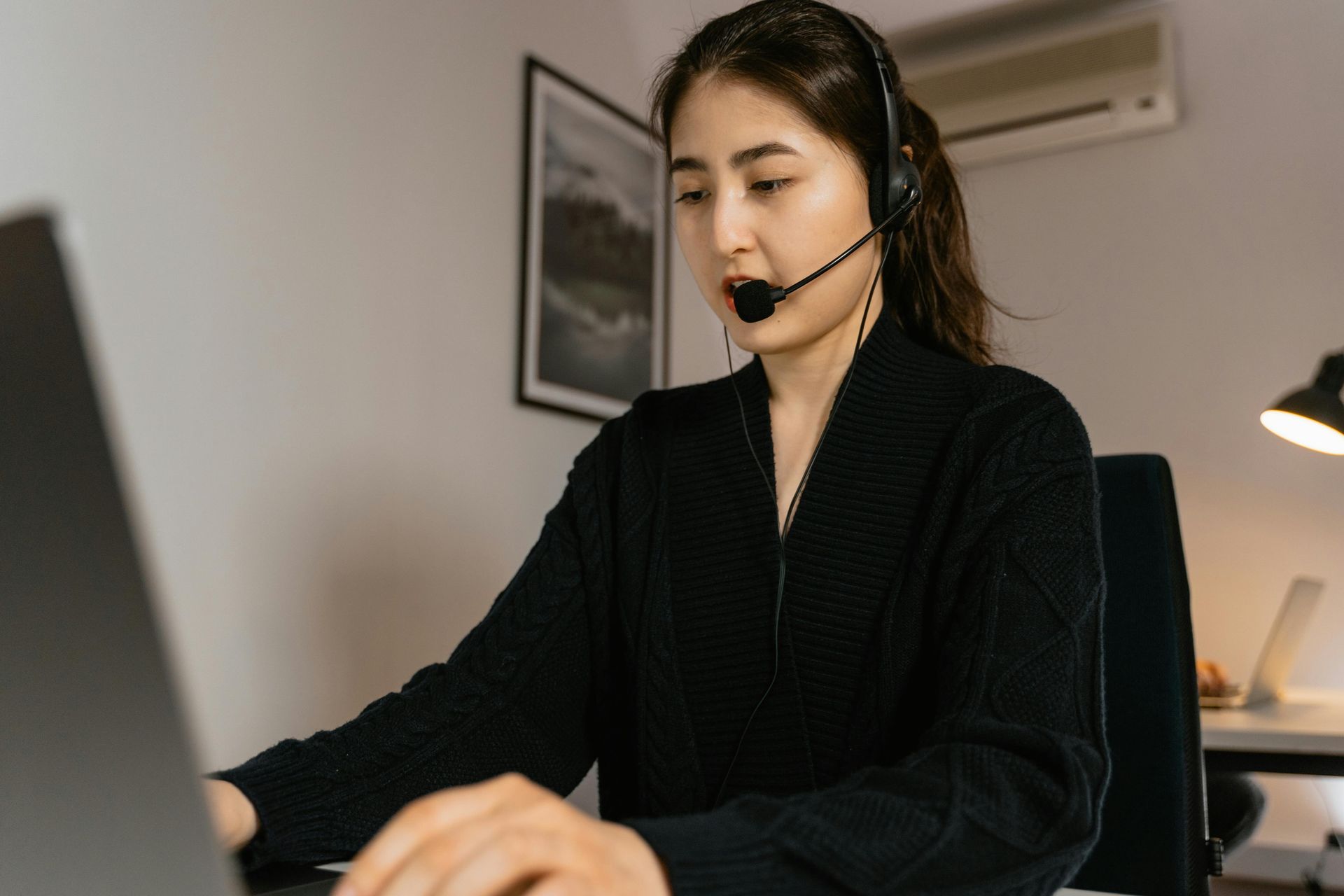 Woman wearing headset works on laptop in office setting.