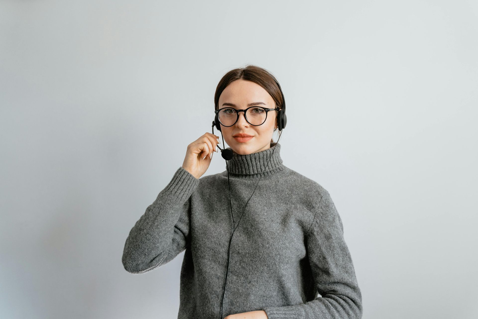 A person wearing a grey sweater, glasses, and a headset, smiling slightly while adjusting the microphone against a wall.