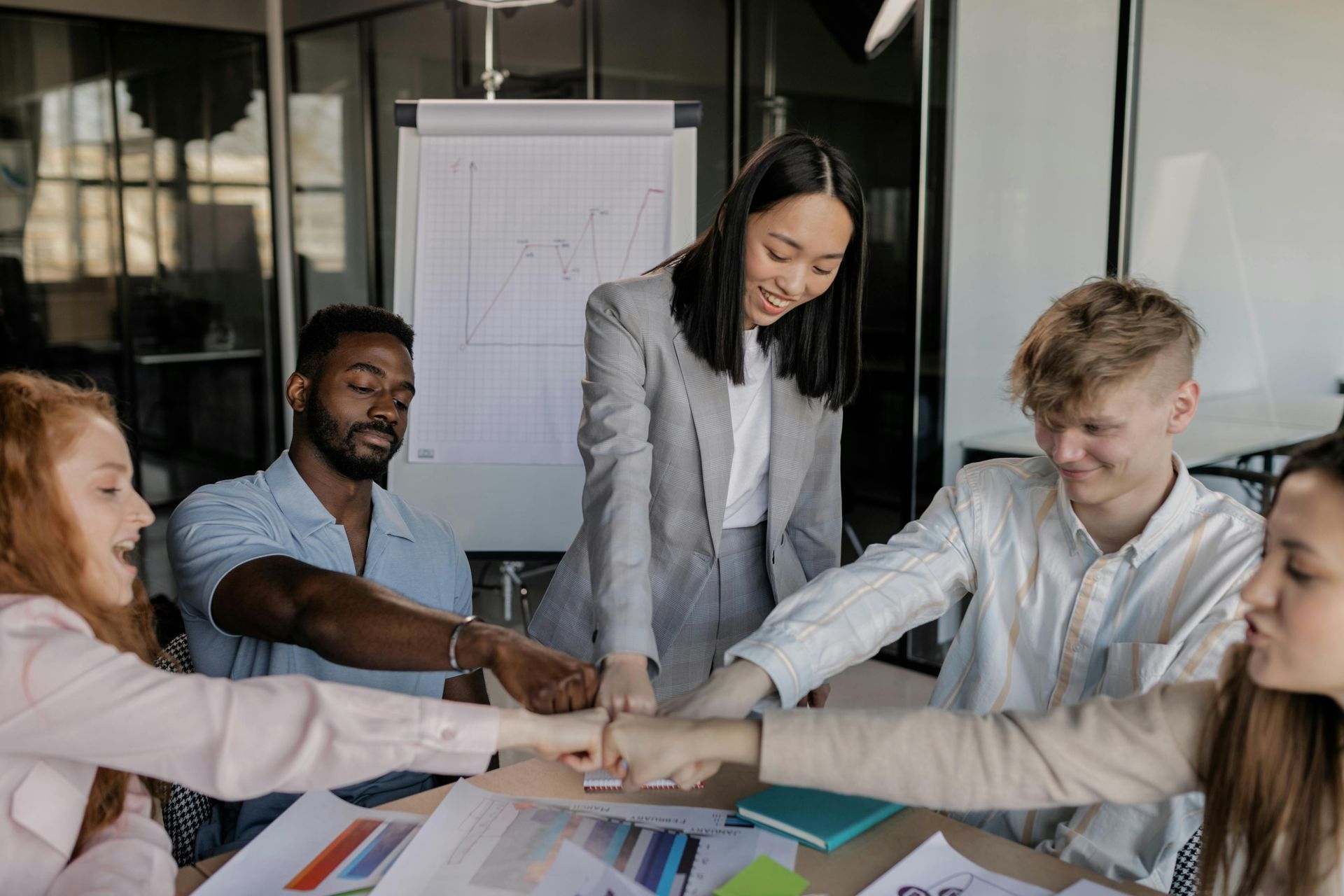 A diverse group of colleagues sit around a table and join hands in a show of unity and collaboration in an office.