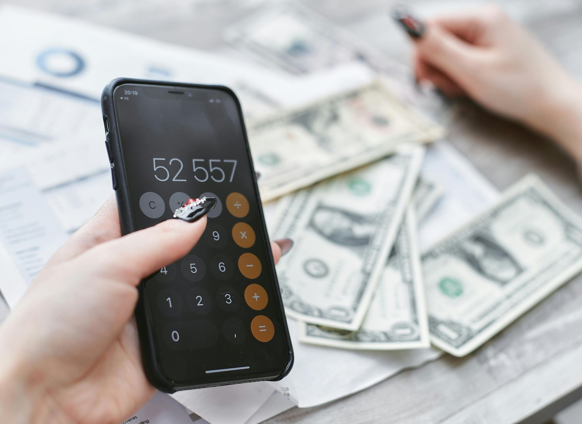 Hand holding a phone displaying calculator; money and documents are on a table.