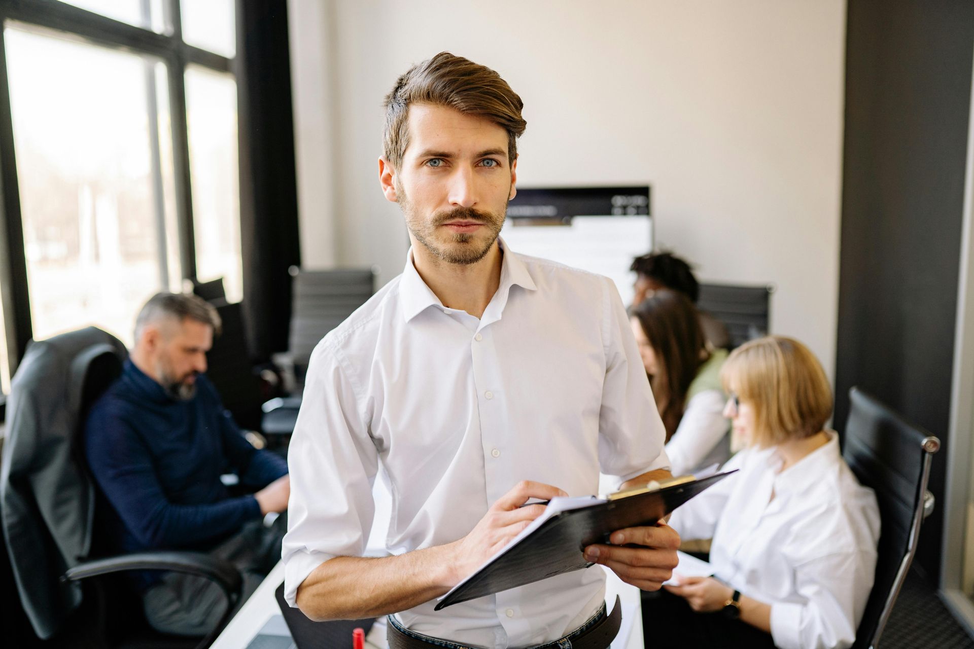 A person holding a folder stands in the foreground of a meeting room, with colleagues working in the background.