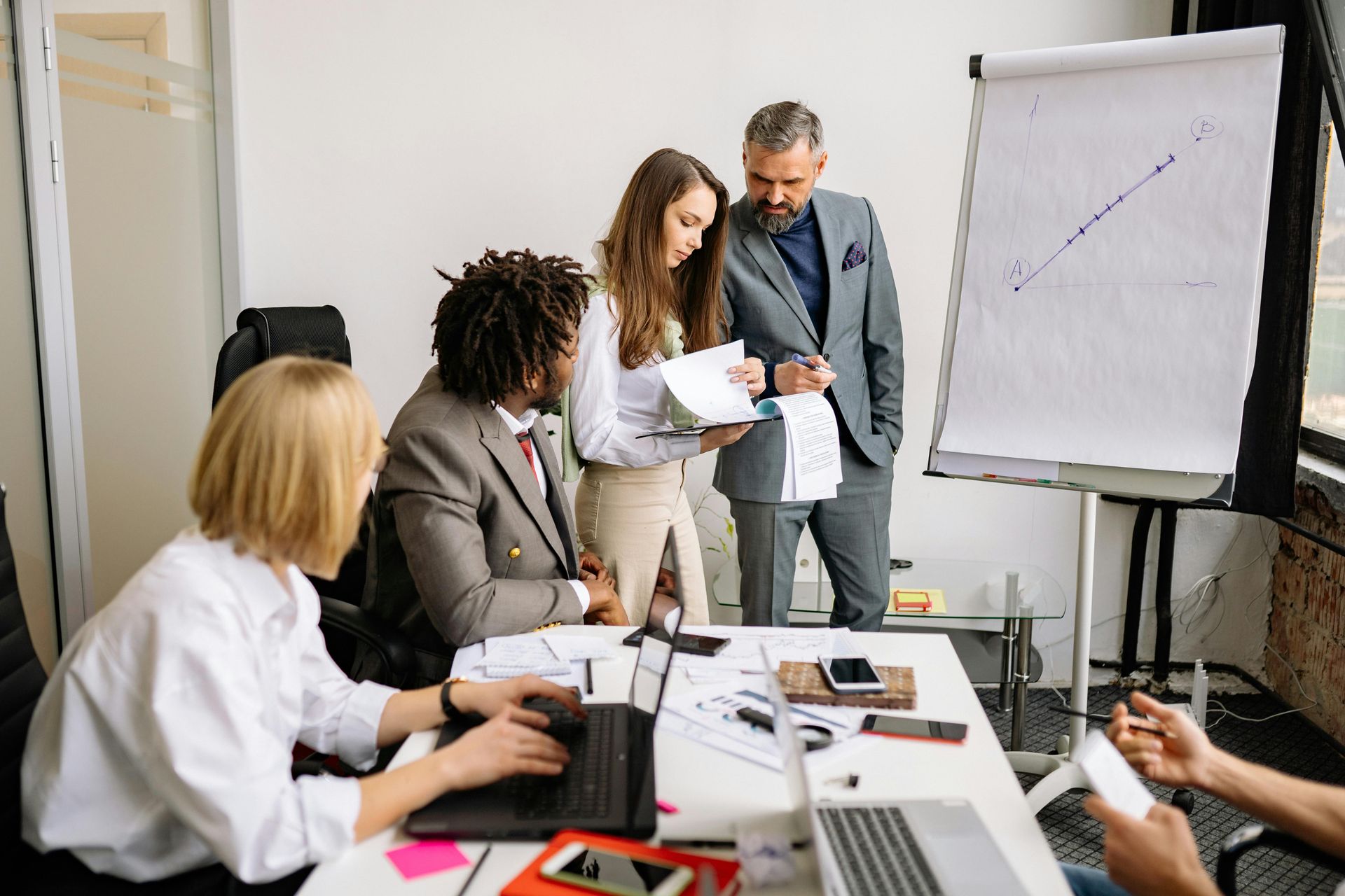 A diverse team collaborates in an office, reviewing documents and a growth chart on a whiteboard during a meeting.