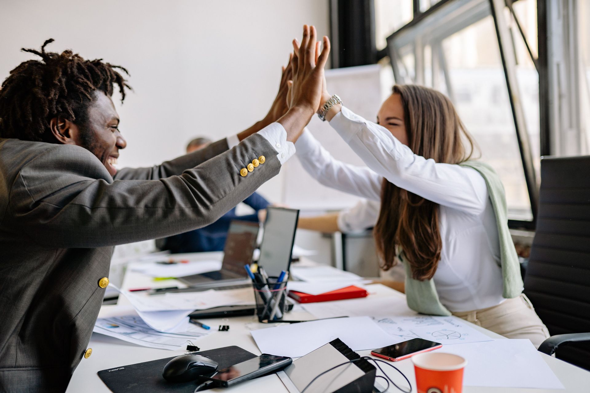 Two coworkers high-five across a desk in a bright office, with laptops and papers scattered around.