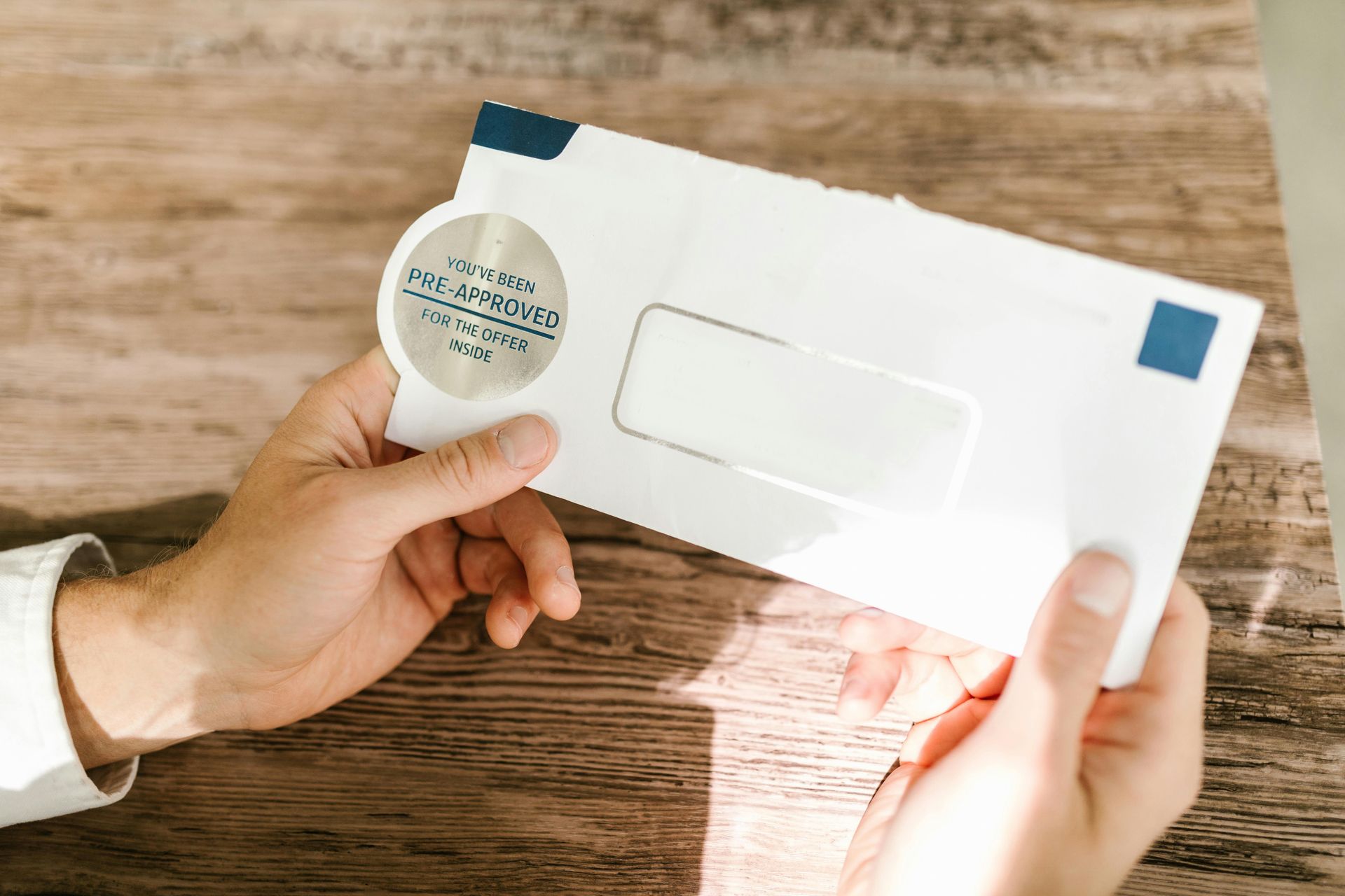 Hands holding a white envelope with a silver circular seal and a windowed address area against a wooden background.
