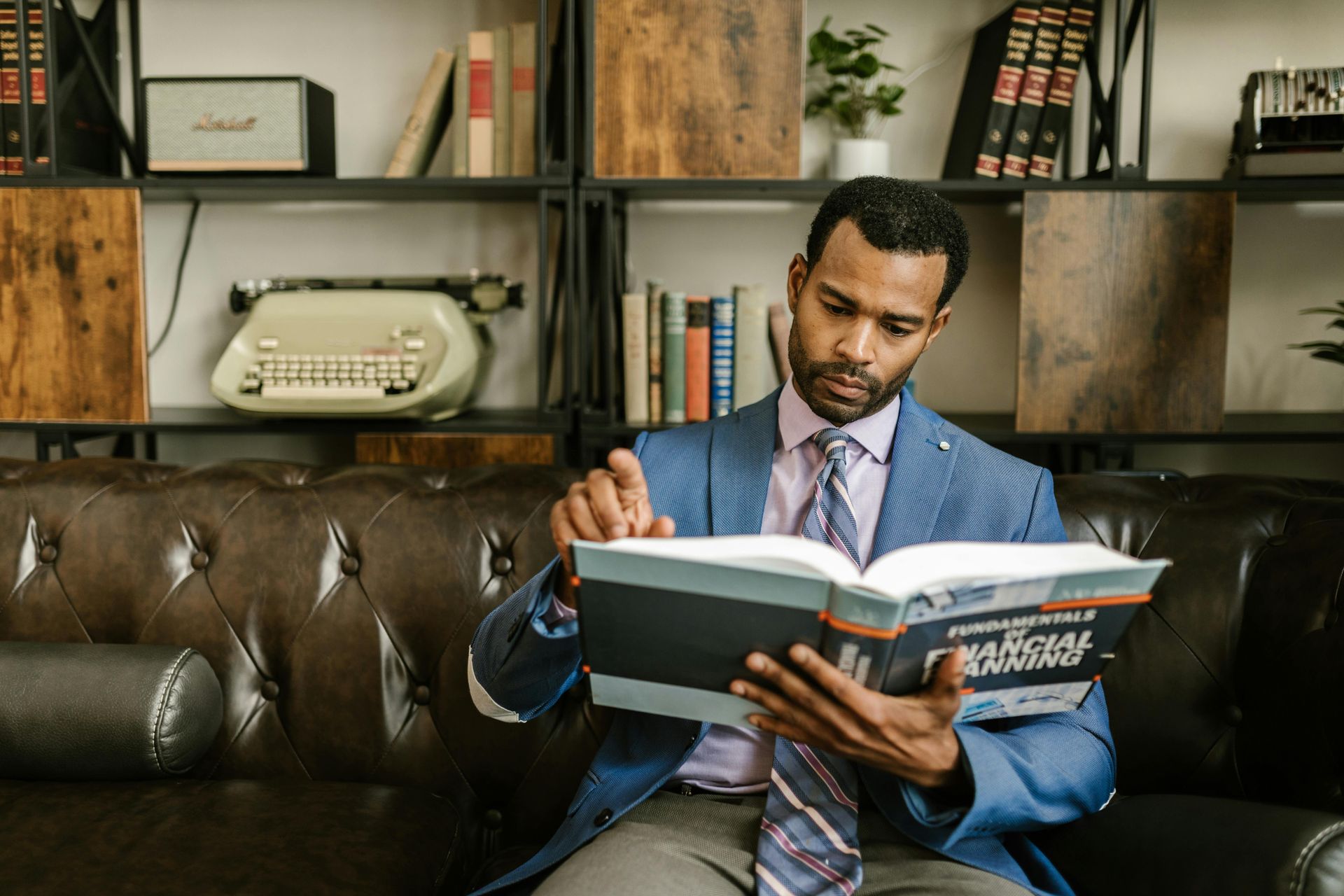 A person in a blue blazer and tie sitting on a leather sofa, reading a book titled 