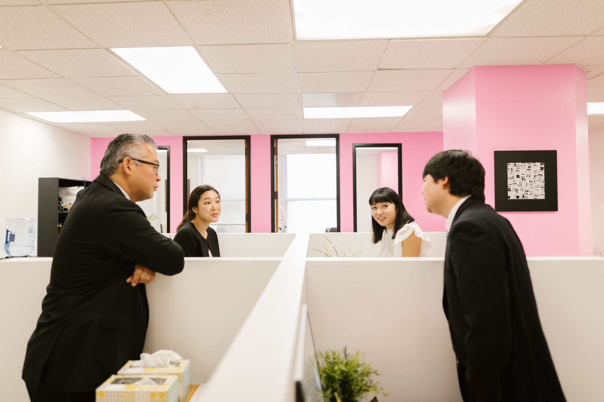 Four people talking across office cubicles in a pink-walled workspace
