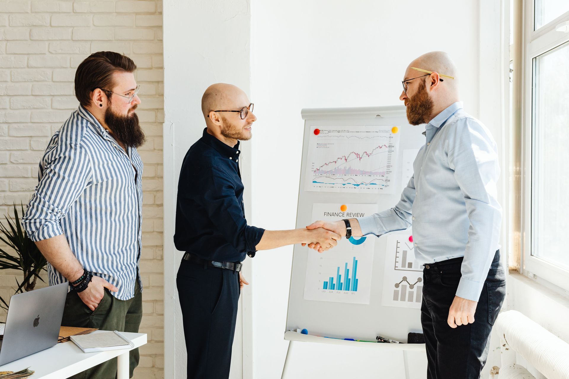 Three coworkers in an office shaking hands beside a flip chart with charts.