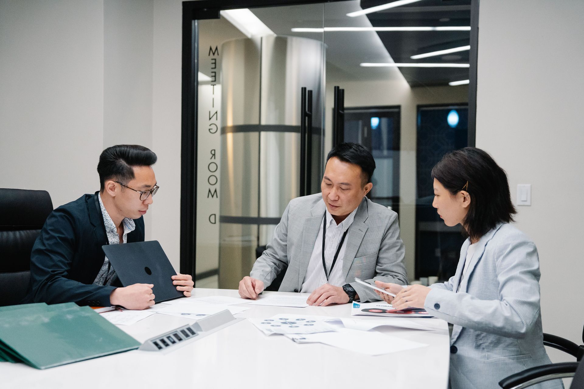 Three coworkers discuss documents around a conference table in a bright office meeting room.