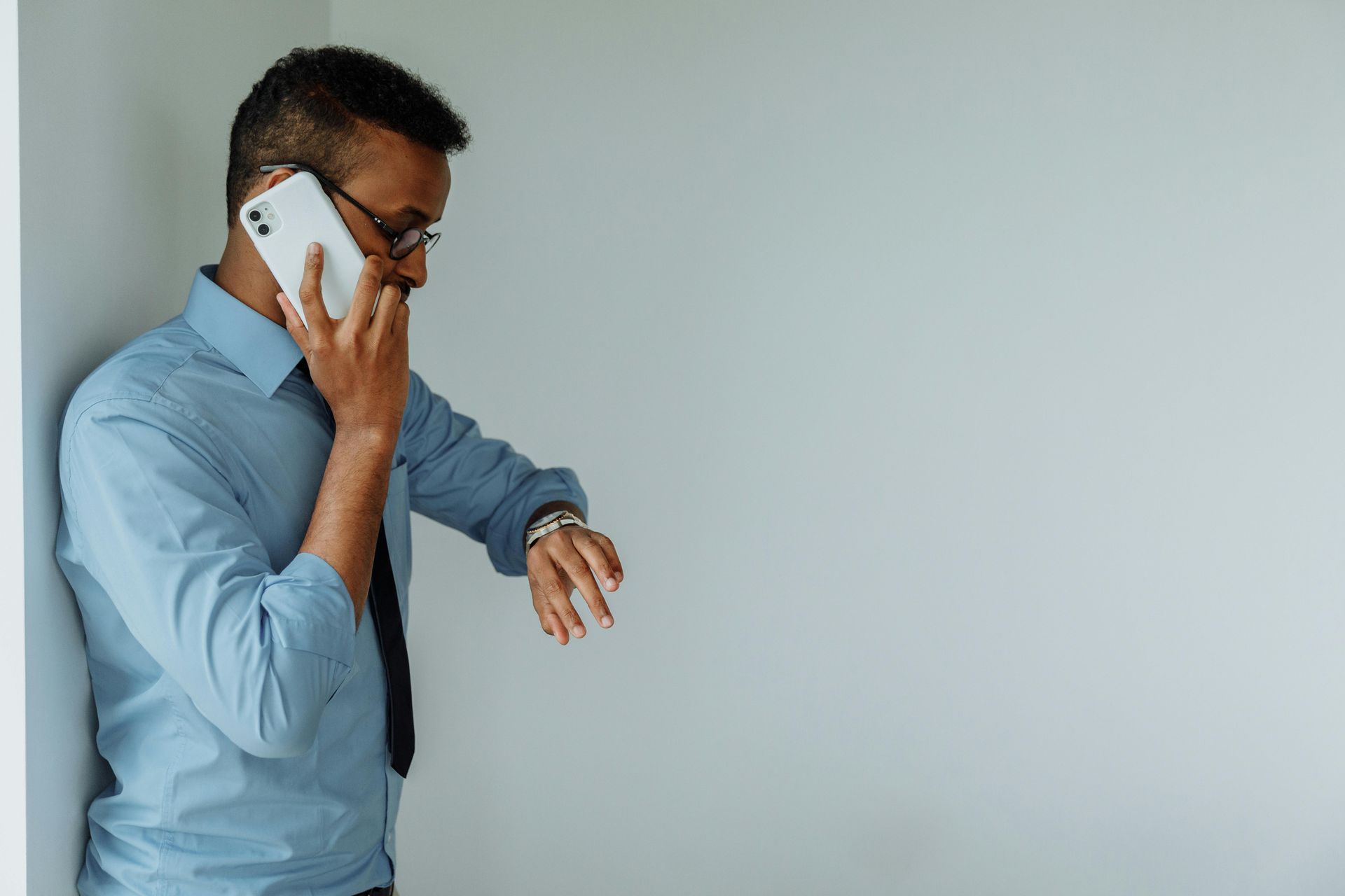 A professional in a blue shirt and tie leans against a wall while talking on a phone and checking a wristwatch.