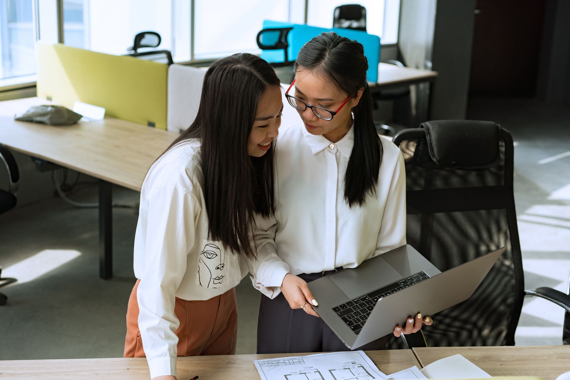Two coworkers reviewing a laptop and printed plans in a bright office