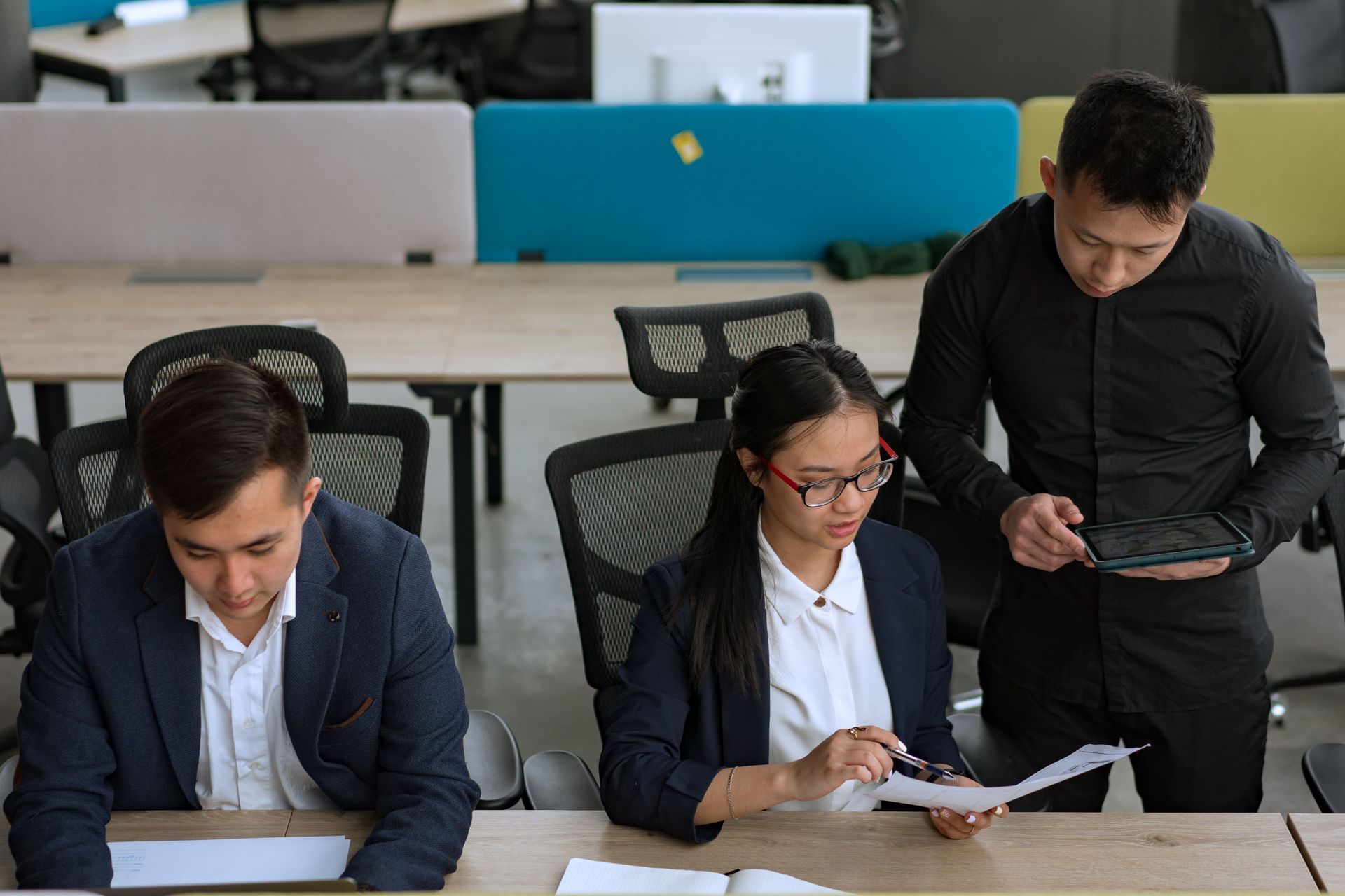 Three people in a modern office, two seated reviewing papers and one standing using a tablet.
