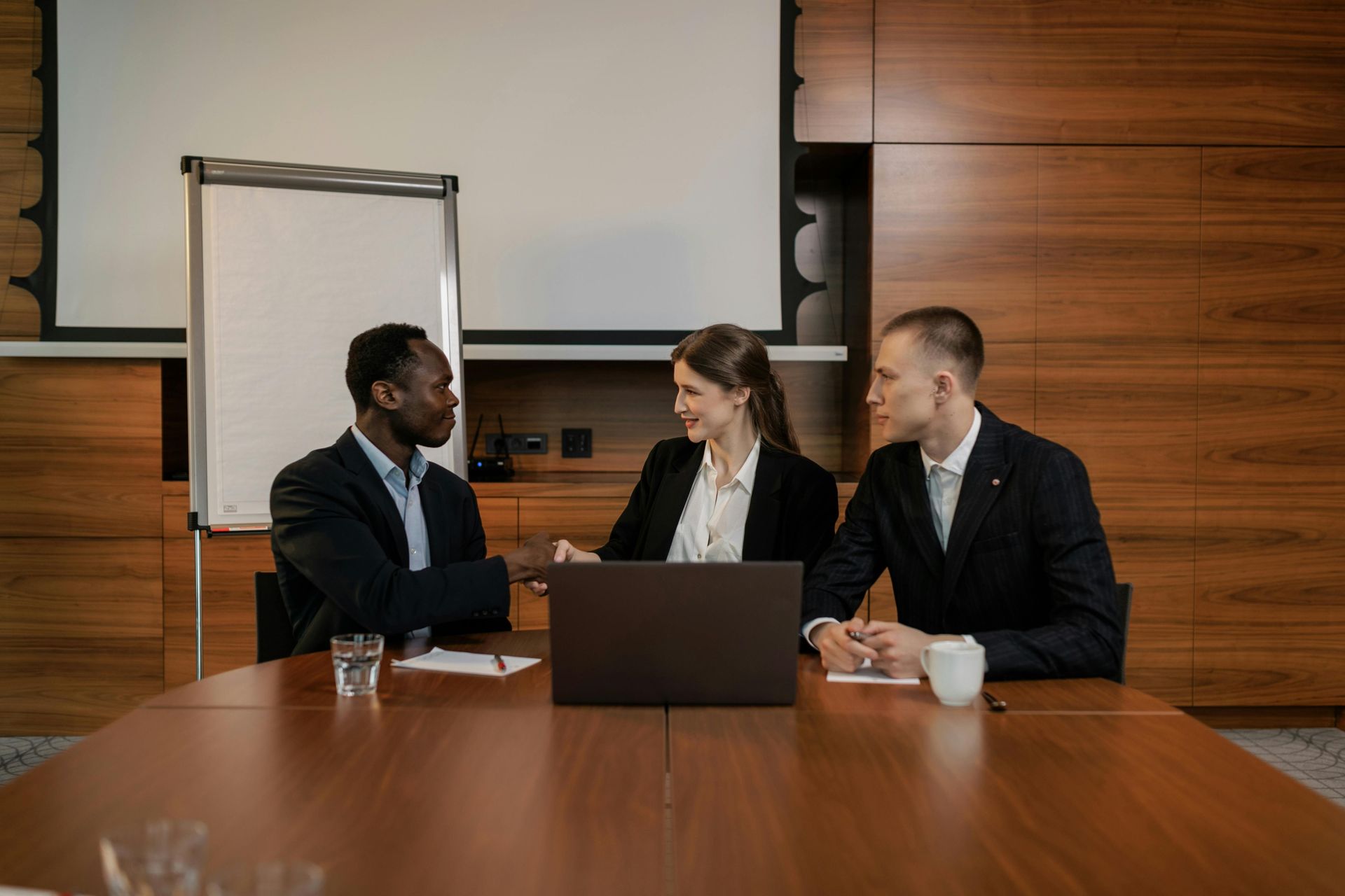 Three colleagues in business attire shake hands across a conference table with a laptop, in a wood-paneled office.
